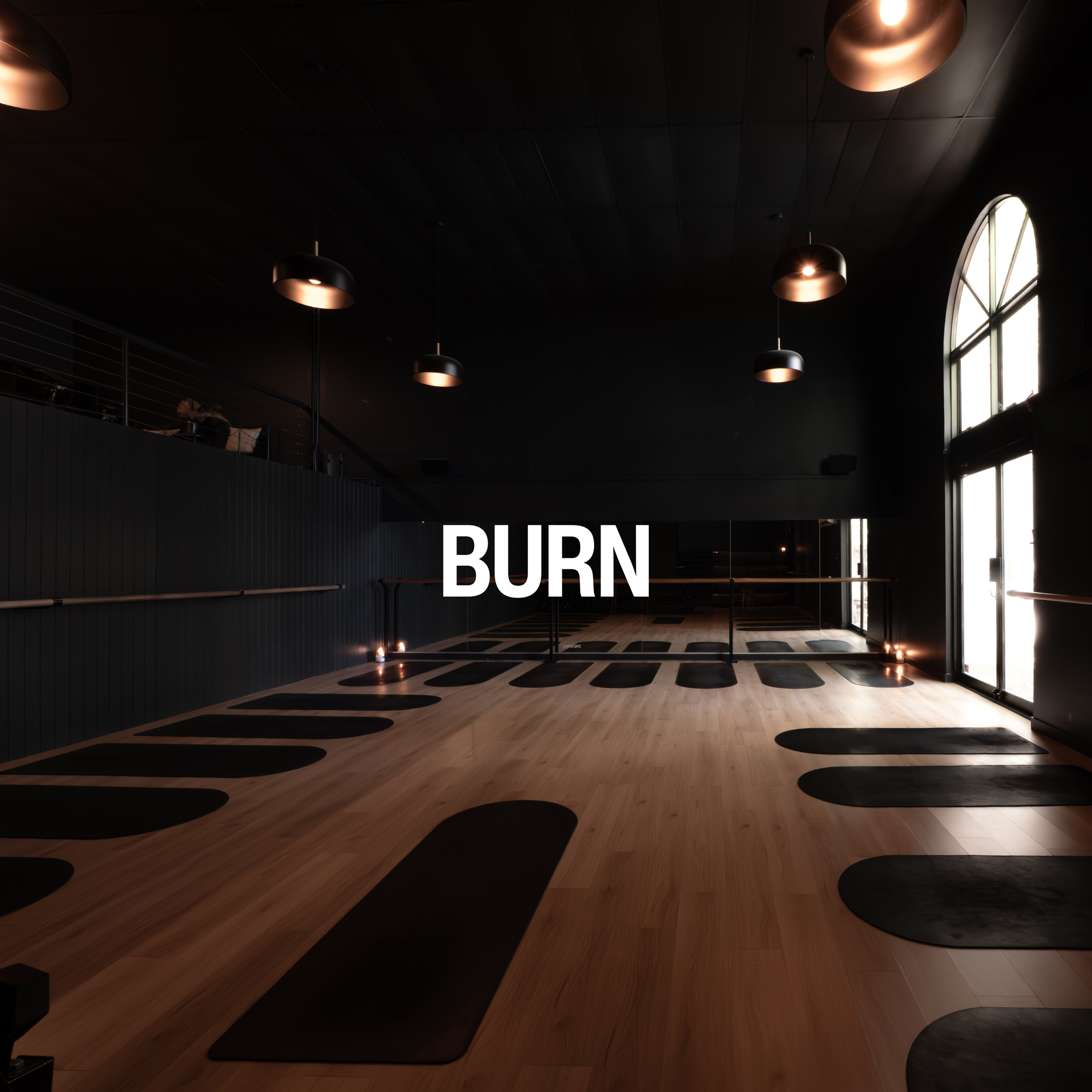 A dimly lit indoor space with a high, black ceiling with hanging pendant lights and large arched window allowing natural light to enter. The floor has a light wood finish with black circular patterns, and there is a sign that reads 'BURN' on a wall.