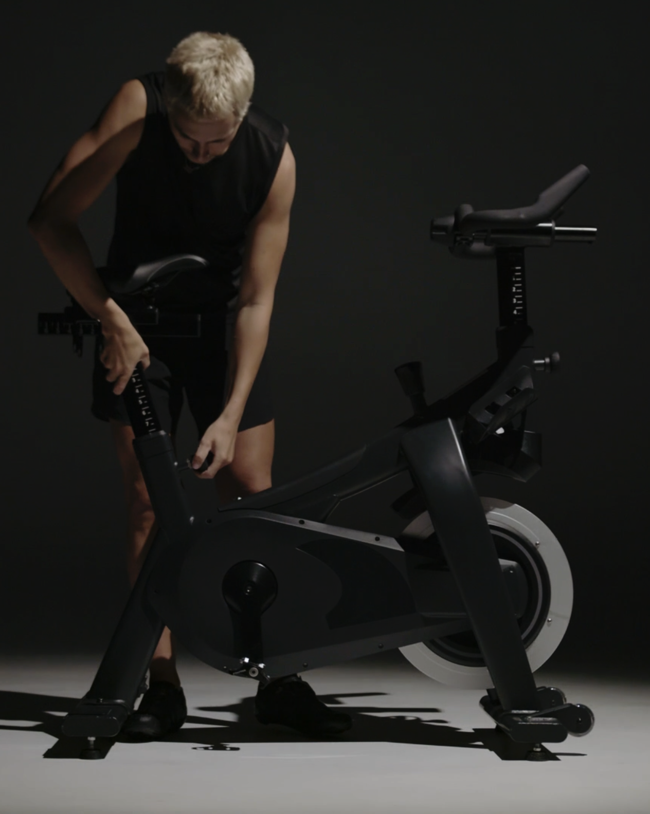 Person with blonde hair adjusting settings on a black stationary exercise bike in a dark room.