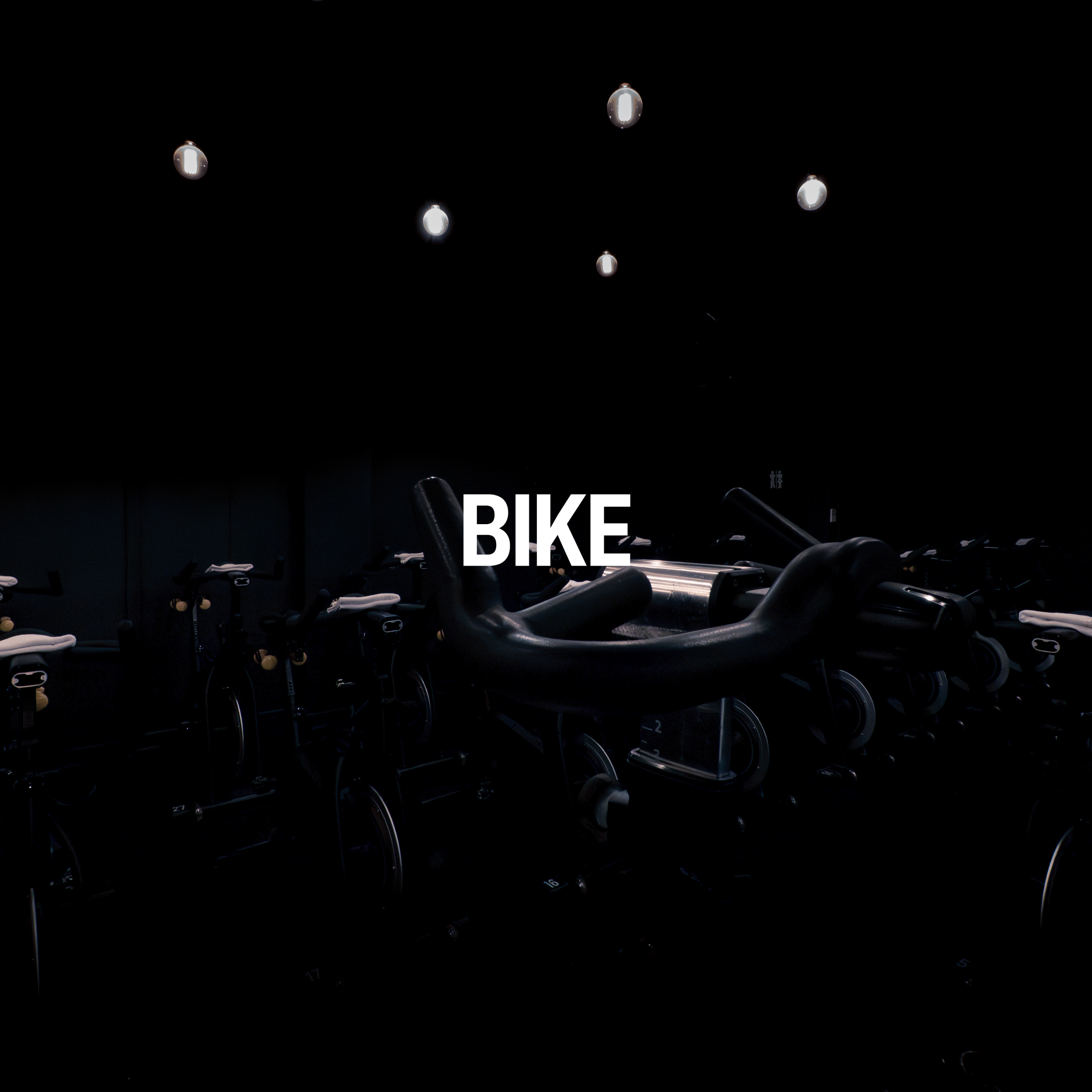 Rows of indoor exercise bikes with a dark background and illuminated lights hanging from the ceiling.