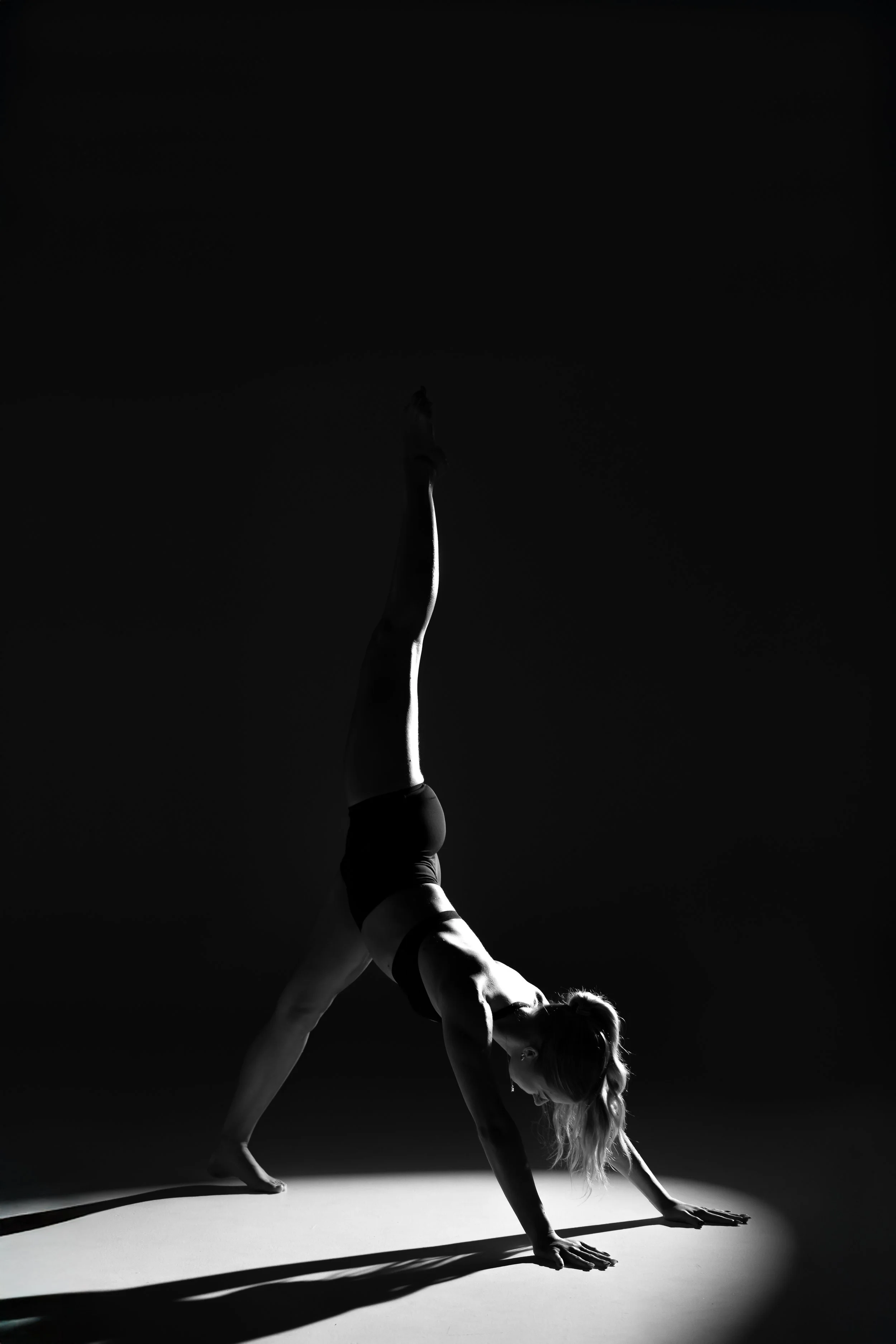 A person performing a yoga pose in a dark setting, with a spotlight creating a sharp shadow on the floor. The person is balanced on their hands with one leg stretched upward, in a handstand variation.