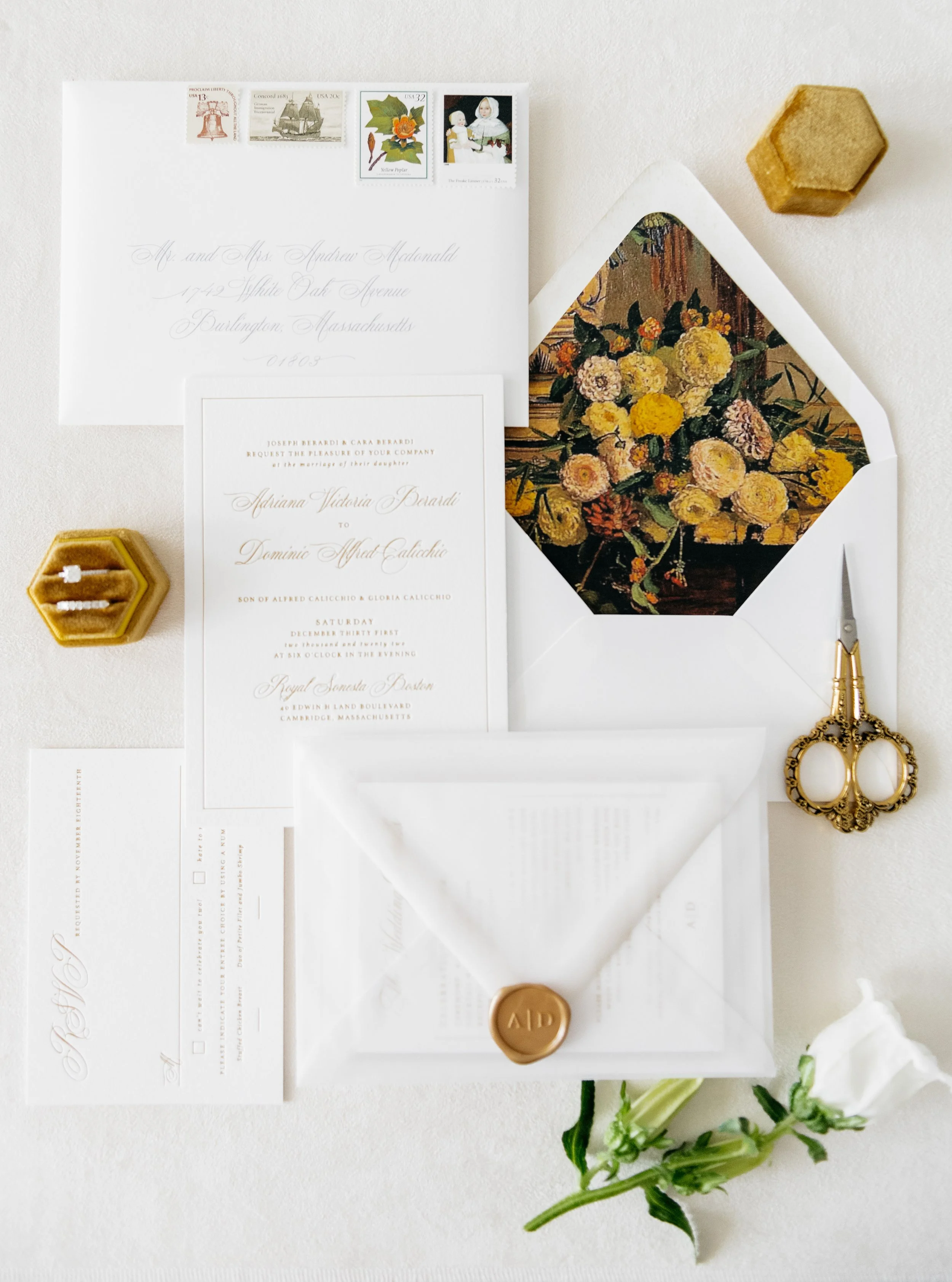 Elegant wedding invitation suite with gold script text, white envelopes, gold wax seal, vintage stamps, floral envelope liner, hexagonal ring box, gold scissors, and a white flower on a white surface. Los Angeles, California 