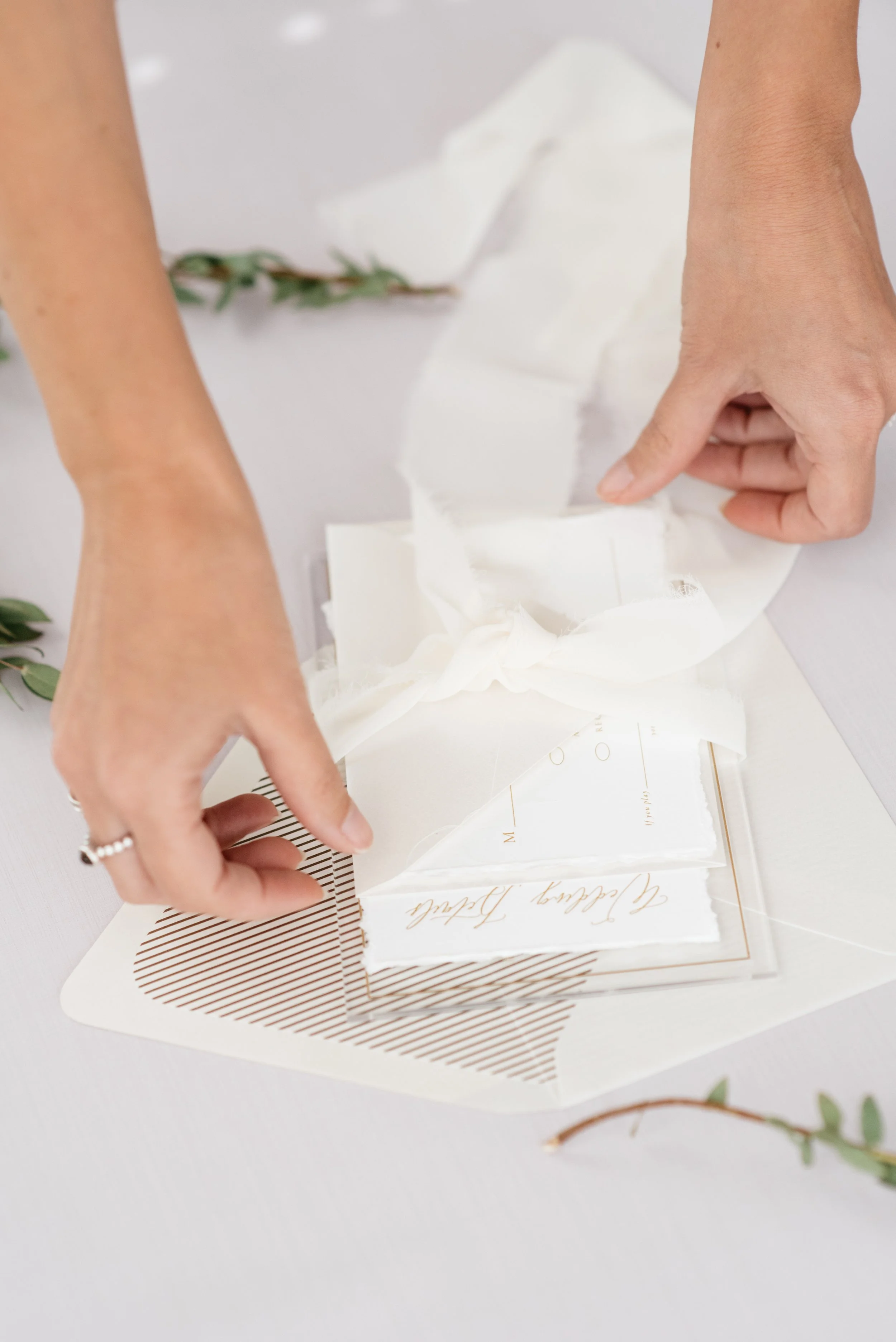 Hands arranging wedding invitations with elegant script, ribbon, and greenery on a white surface. Charleston, South Carolina 