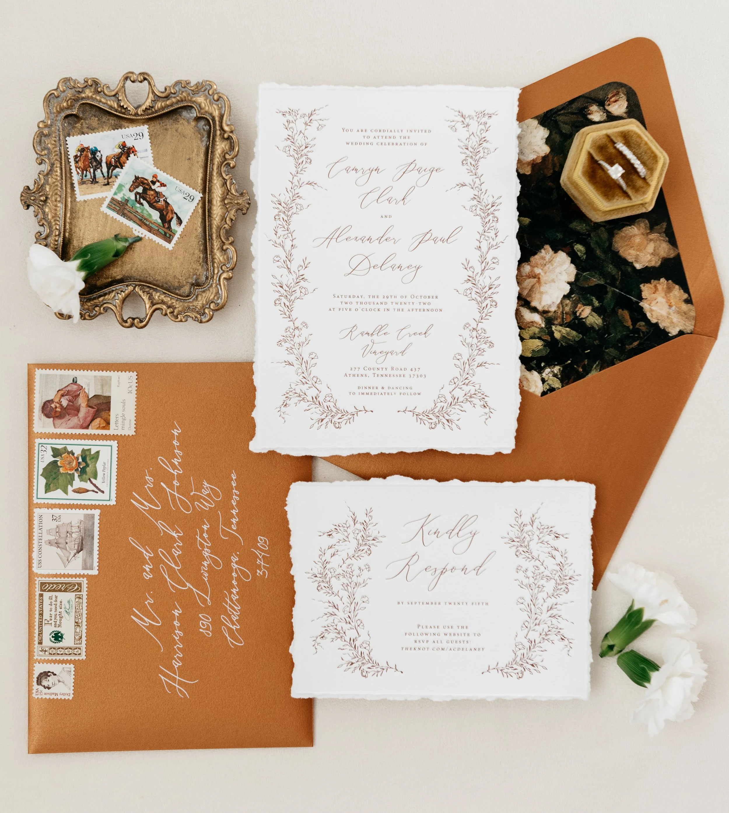 Vintage-style wedding invitation set with floral designs, ornate tray with stamps, brown envelopes, and white flowers