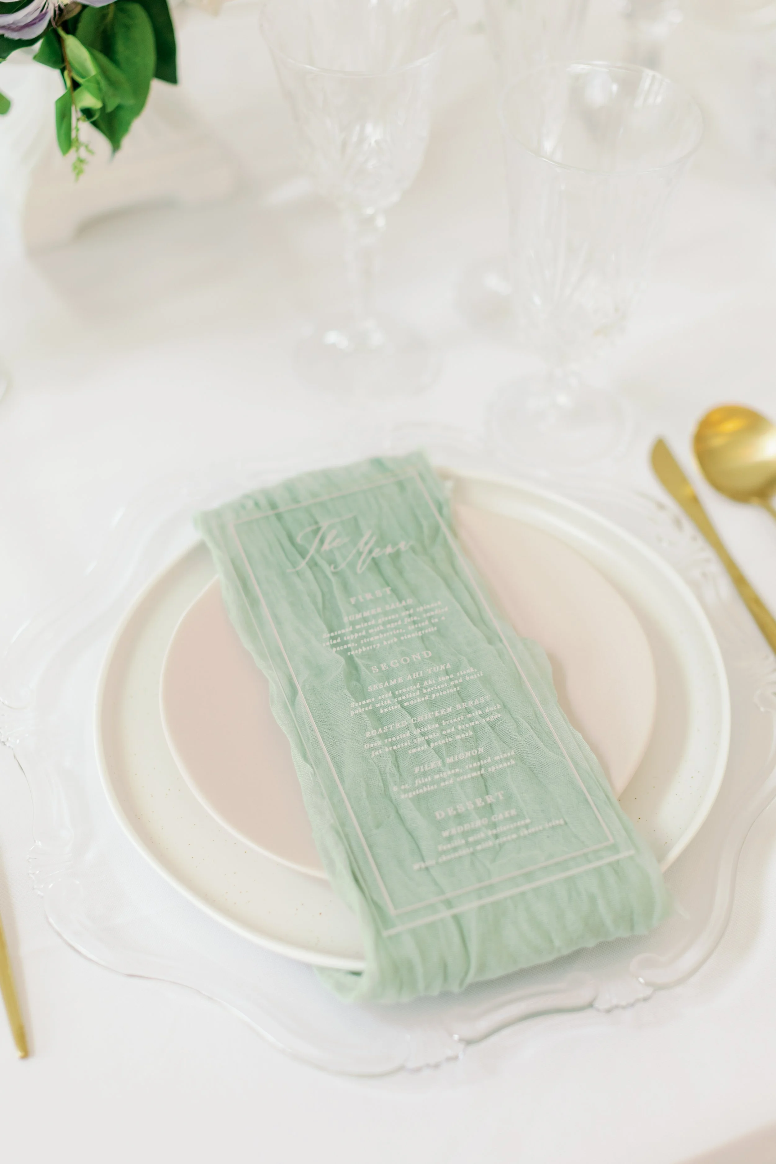 A formal dining table setting with white plates, a green fabric menu, crystal glasses, and gold cutlery on a white tablecloth.