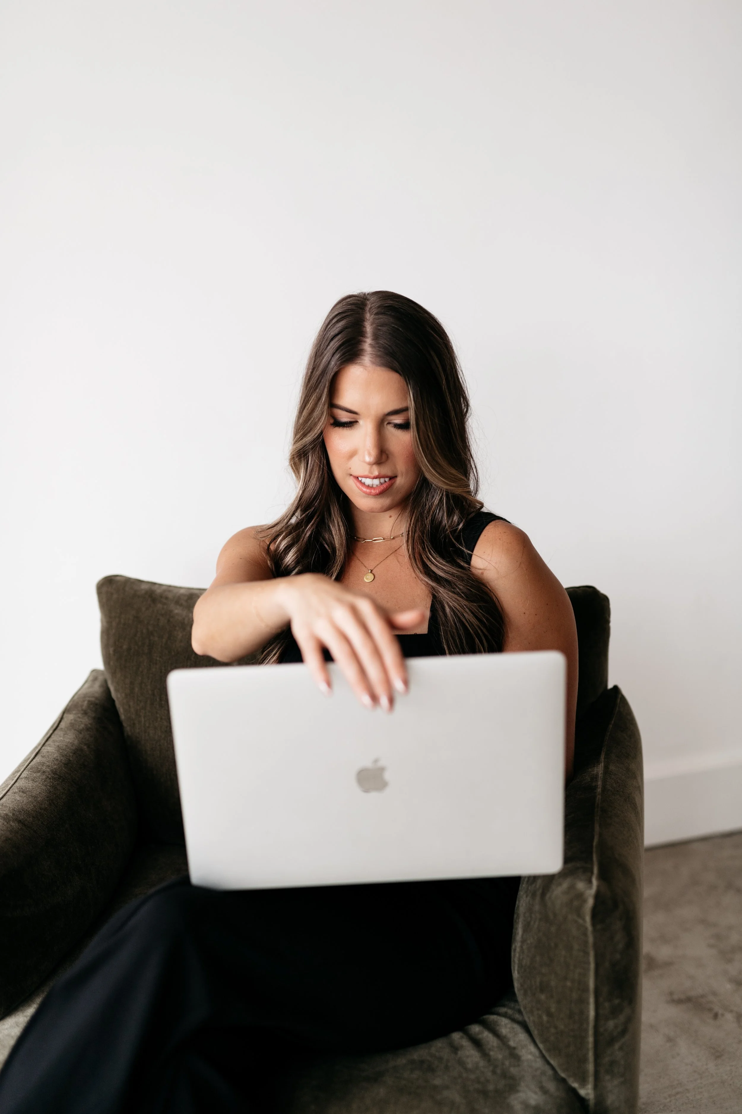 Woman with long brown hair sitting on a dark sofa, looking down at an open silver MacBook on her lap, with a neutral plain white wall in the background.