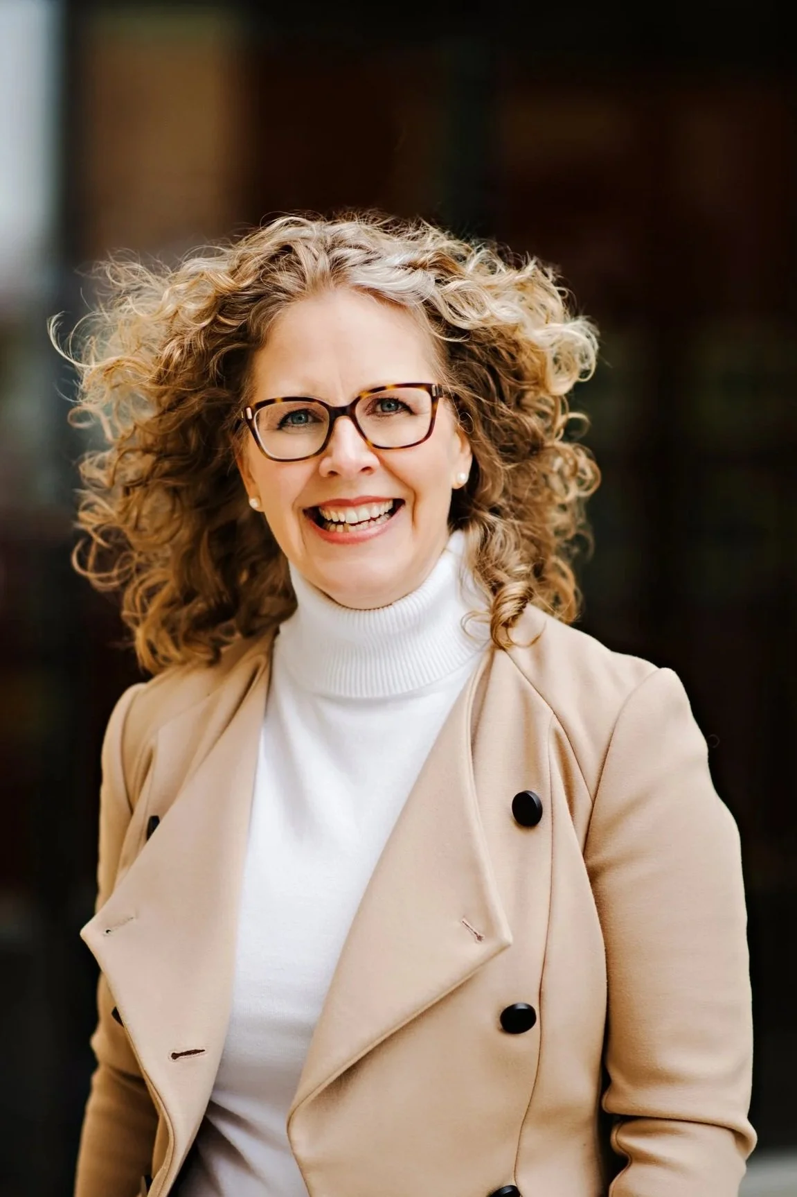 A woman with curly blonde hair, glasses, and a bright smile, wearing a beige jacket over a white turtleneck, outdoors.