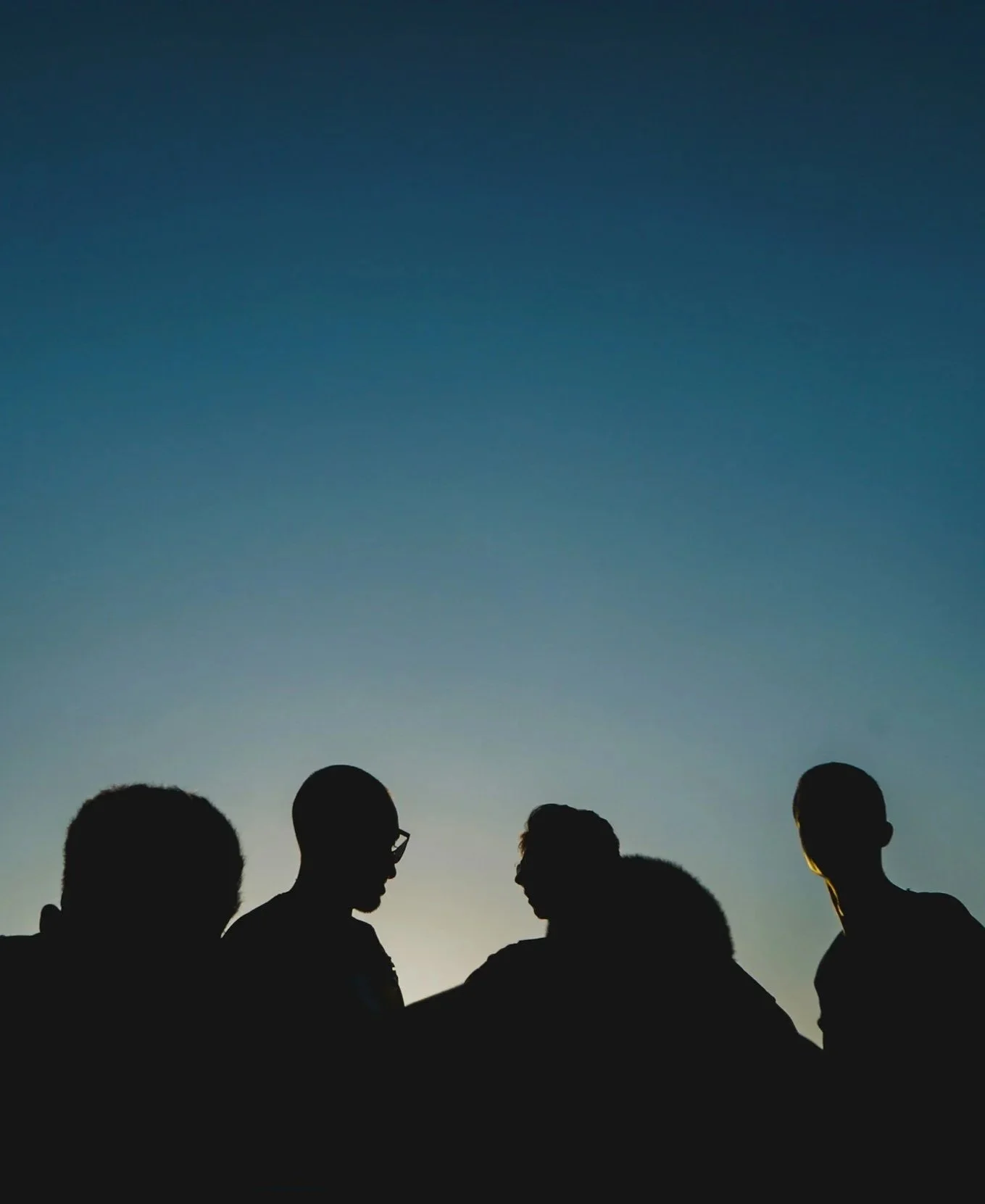 Silhouettes of a group of people standing outdoors during sunset or dusk with a dark blue sky in the background.