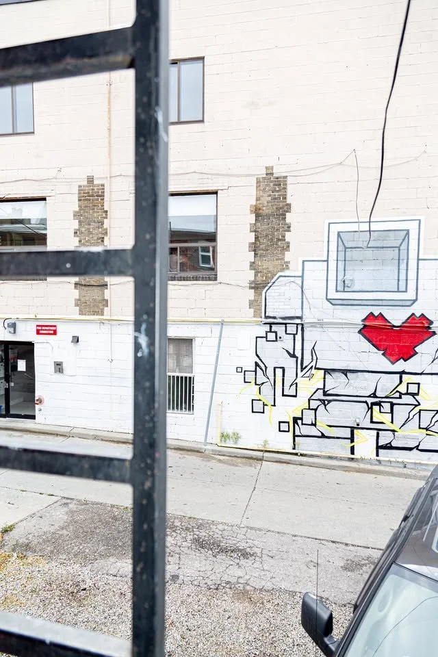 Graffiti on a white brick building wall, including a pixelated red broken heart, black and yellow abstract shapes, and some geometric lines, with part of a metal fence and a vehicle in the foreground.