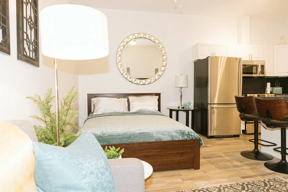 A cozy studio apartment with a bed, nightstand, lamp, mirror, kitchen area, refrigerator, microwave, and two bar stools.