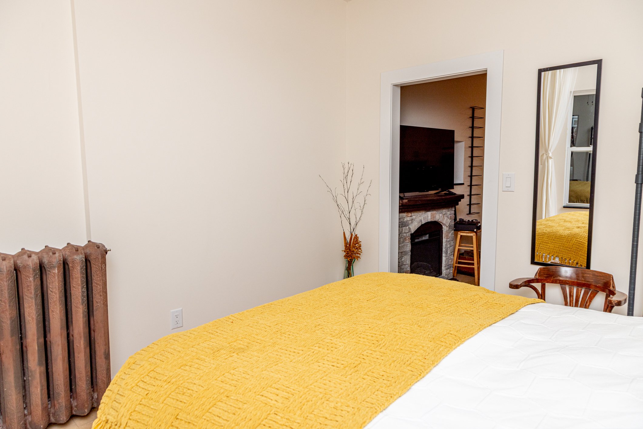 A bedroom with a yellow quilt on the bed, a vintage radiator on the left, a tall mirror on the right, and a fire place with a TV above it in the background.