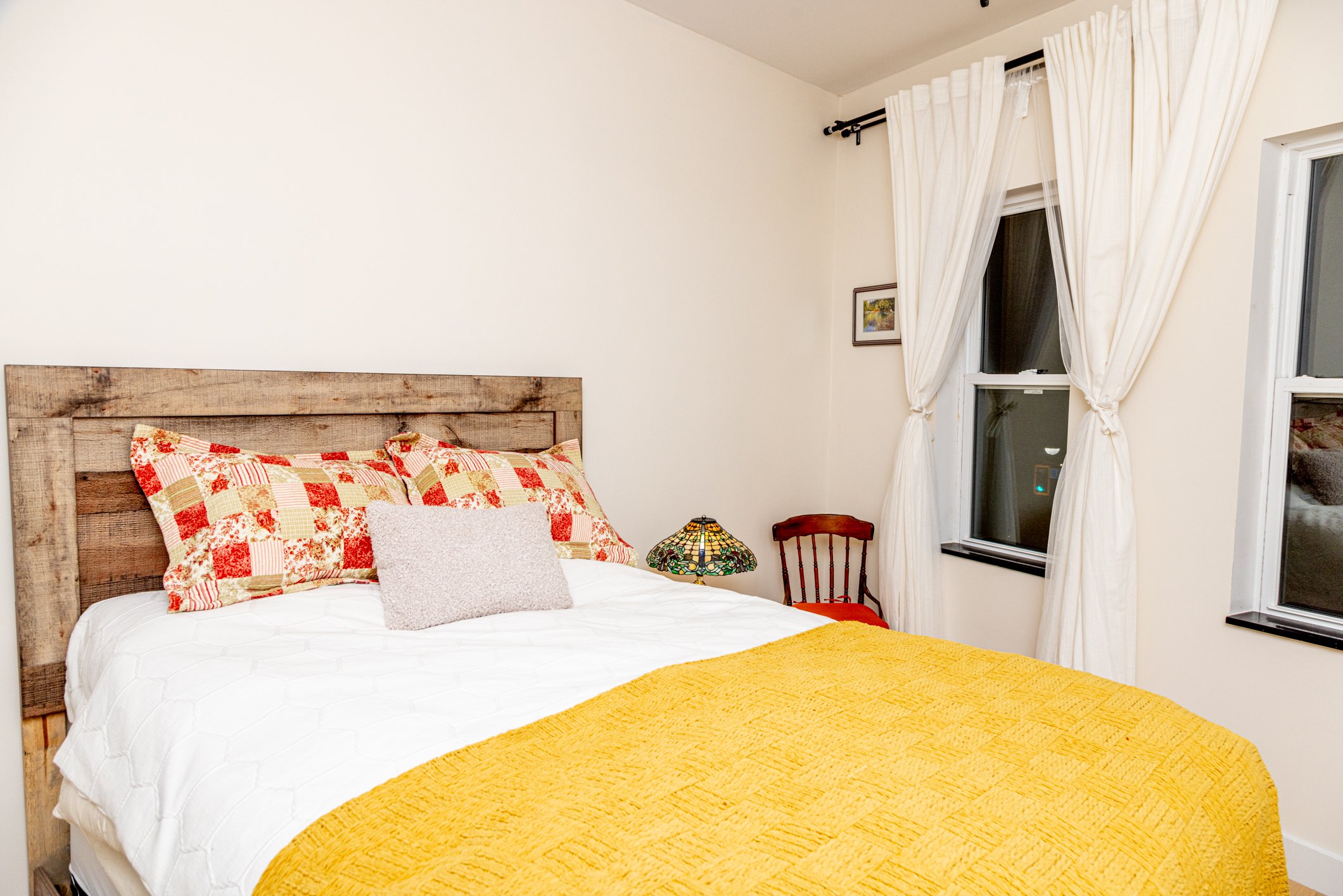 A cozy bedroom with a rustic wooden headboard, a bed with quilted pillows, a stained glass lamp, and cream-colored curtains next to two windows.