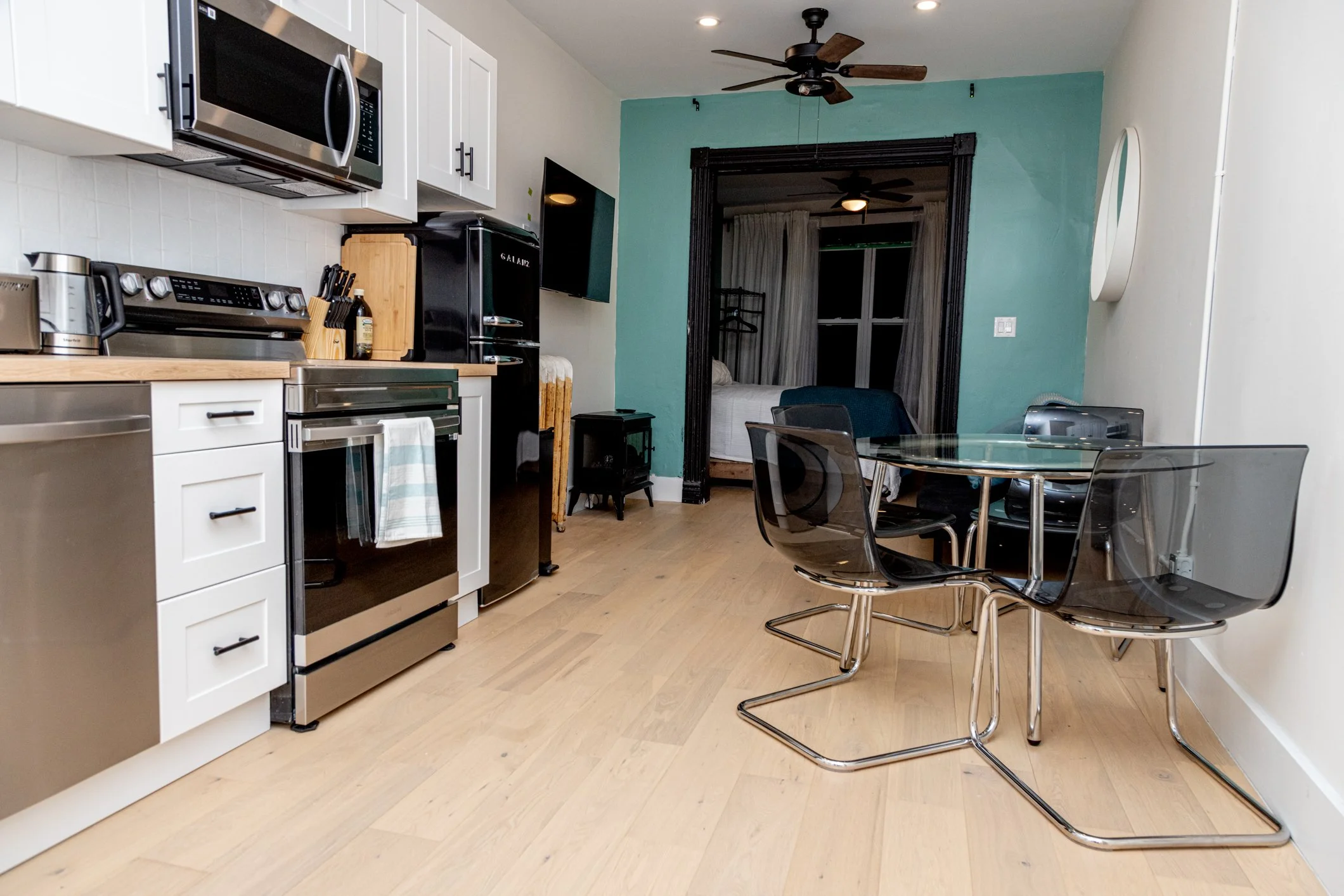 A kitchen with white cabinets, stainless steel appliances, a round glass dining table, and black modern chairs. There is a bedroom visible through a black-framed doorway with teal walls and white curtains.