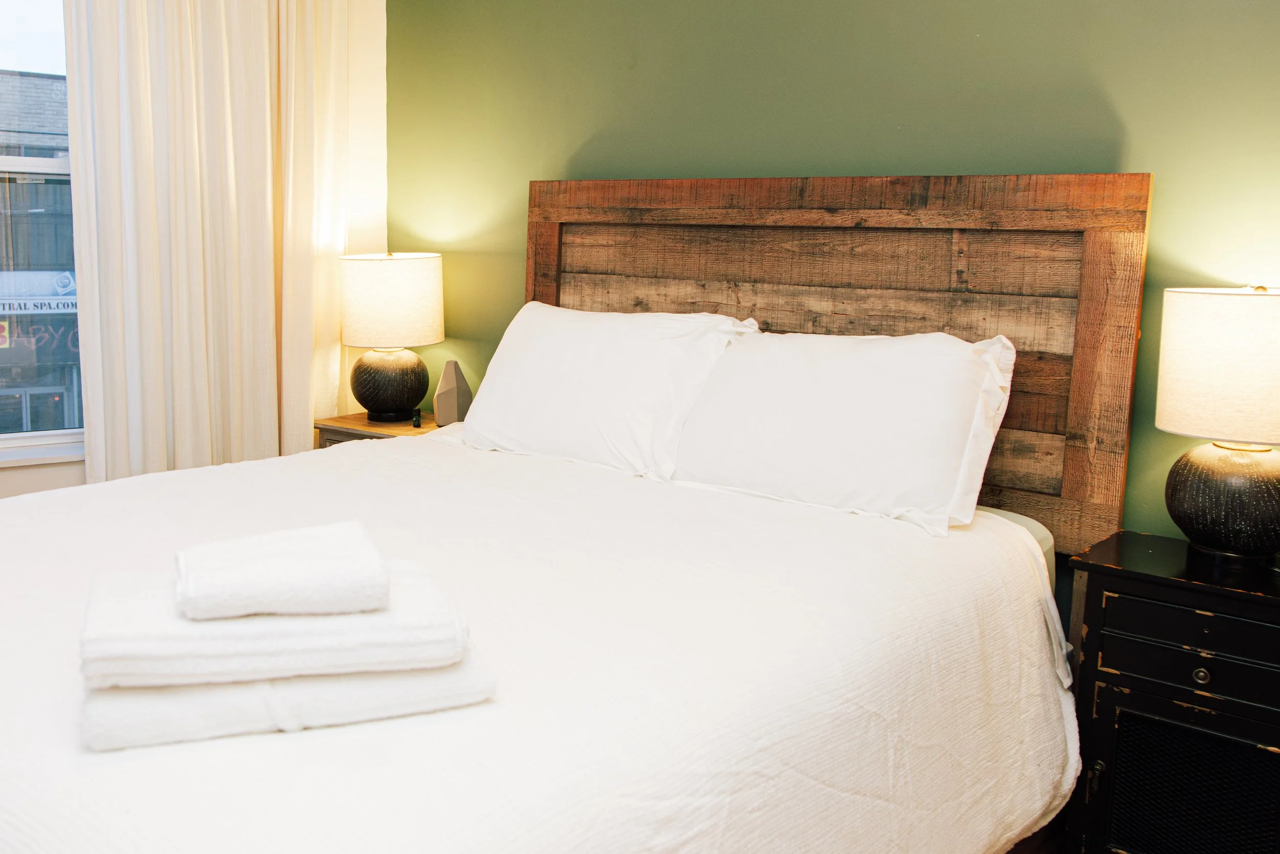A neatly made bed with white linens, two folded towels, and pillows in a bedroom with a rustic wooden headboard, bedside lamps, and green walls.