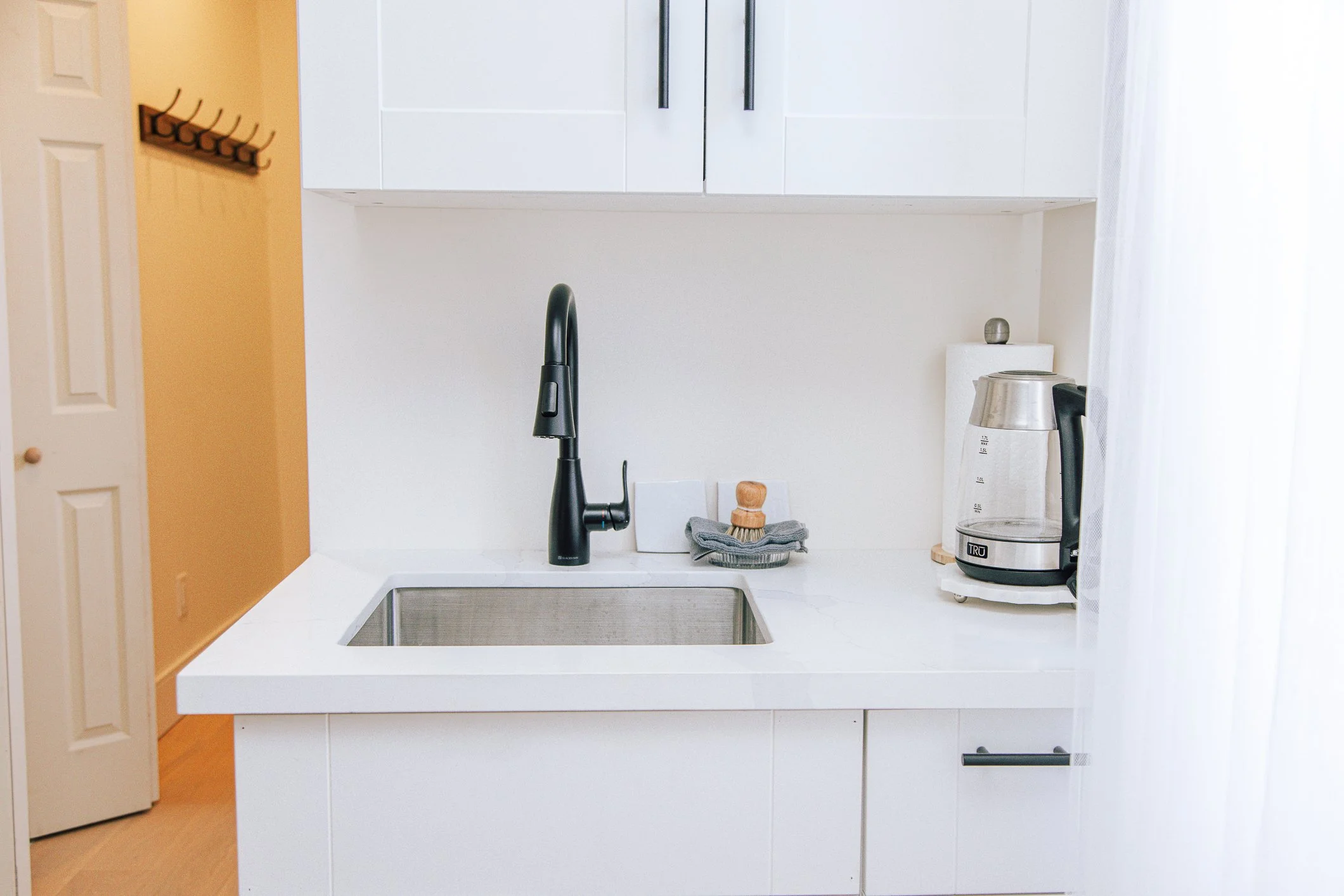 A modern kitchen with a white countertop, black faucet, and a coffee maker on the right side. There is a yellow wall on the left, a white cabinet above the sink, and a white curtain on the right side.