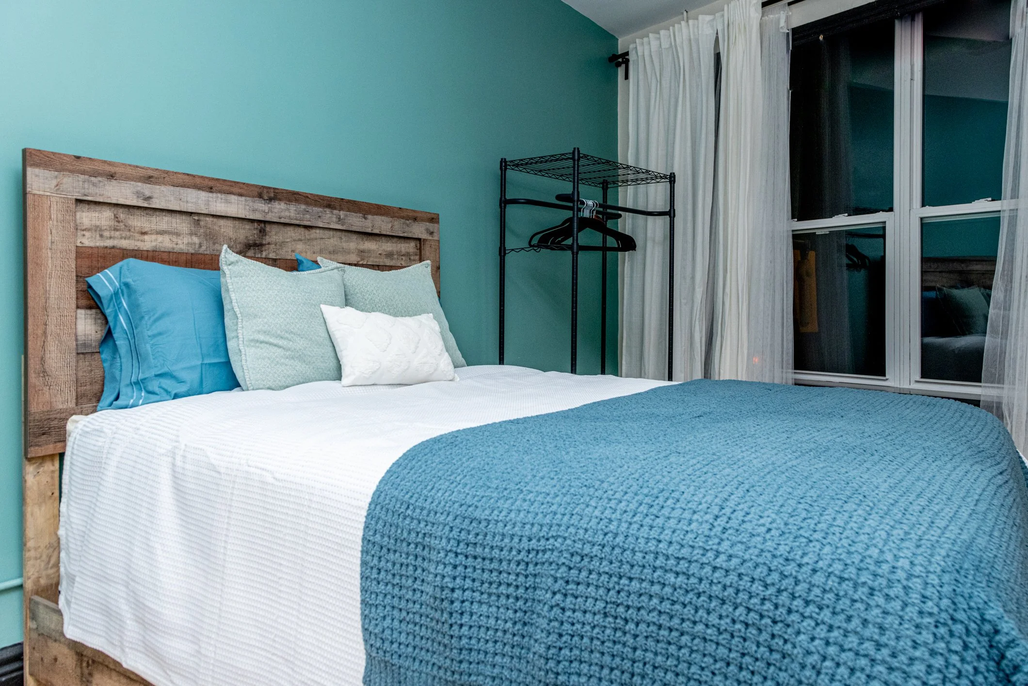 A bedroom with a rustic wooden headboard, white bedding, and blue and mint green pillows, next to a large window with sheer white curtains and a black metal clothing rack.