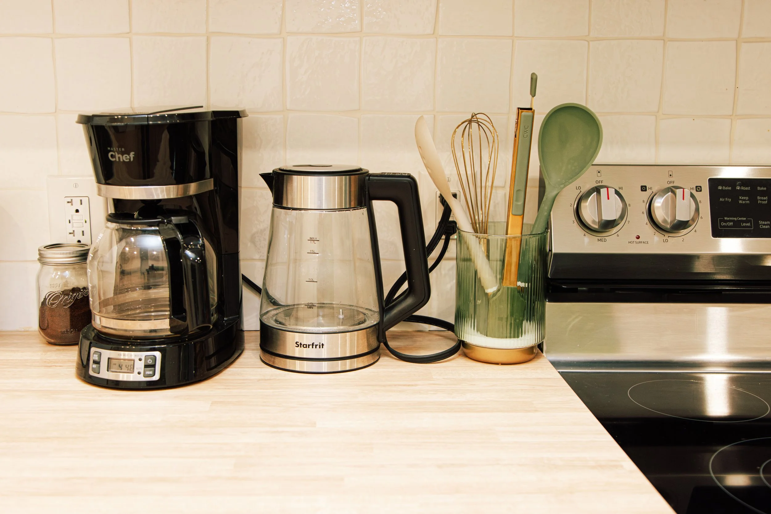 Kitchen countertop with coffee maker, glass coffee pot, electric kettle, utensils in a green glass container, and a stove in the background.