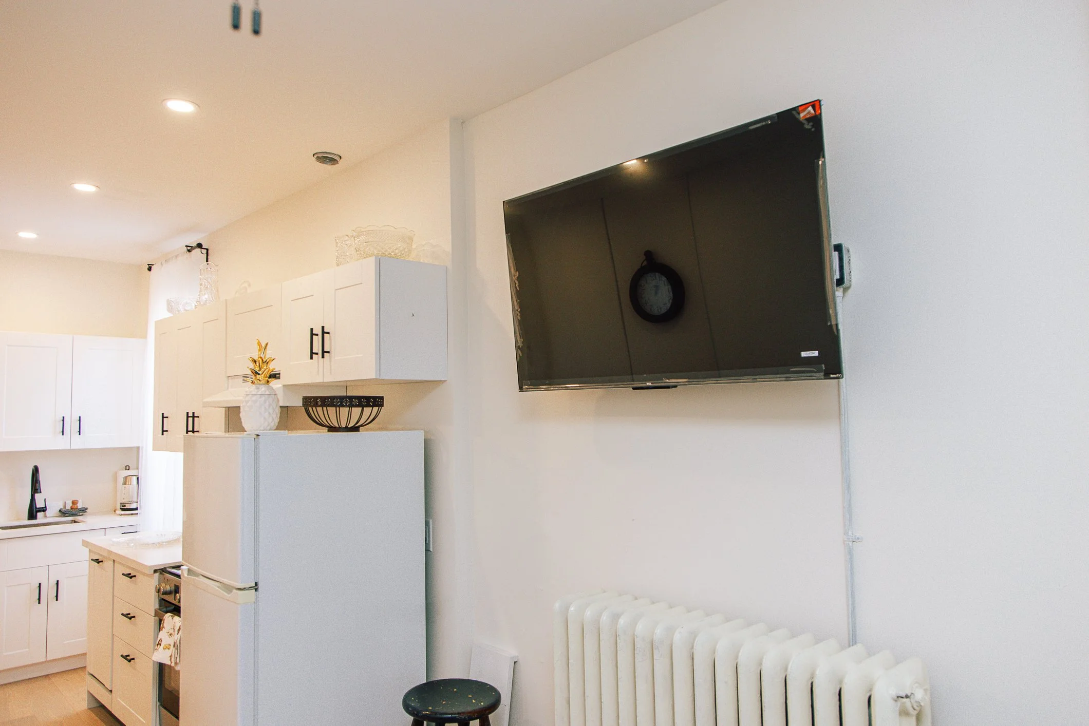 A living space with white cabinets, a white refrigerator, a small stove, a black stool, a radiator, and a wall-mounted flat-screen TV. Decor includes a pineapple-shaped vase and a black wire bowl.