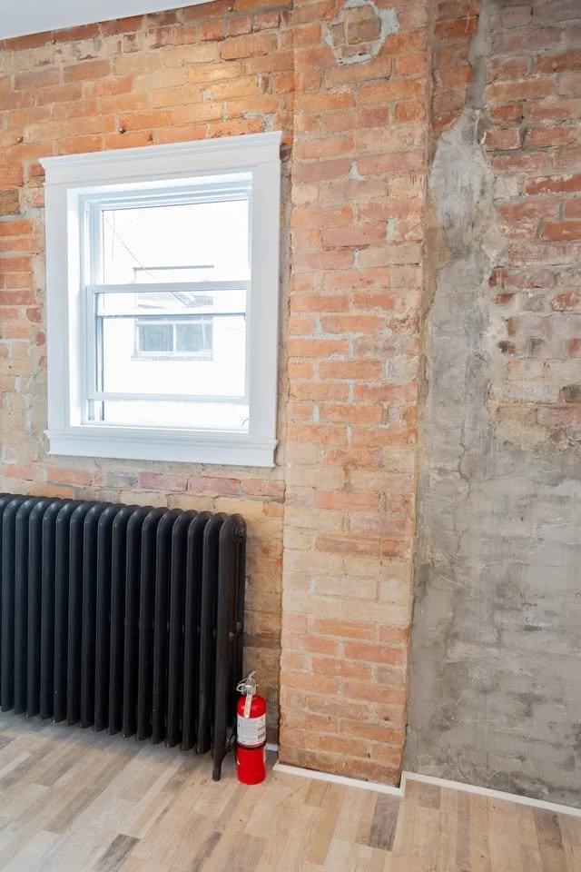 Interior of a room with exposed brick wall, a white-framed window, a black radiator, and a red fire extinguisher on the wooden floor.
