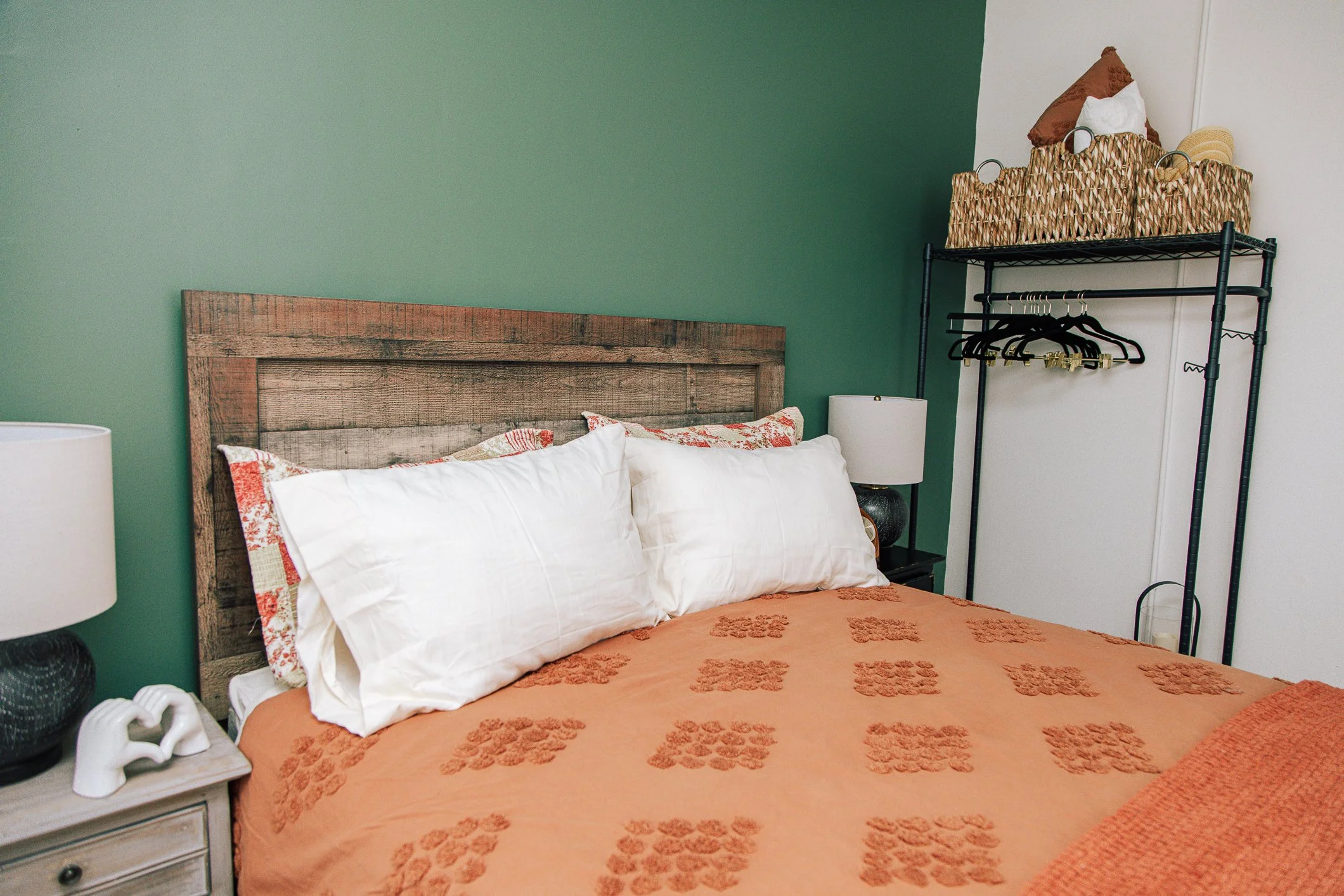 A bedroom with a rustic wooden headboard, green accent wall, and bed with white pillows and an orange bedspread with floral patterns. A black metal shelving unit holds woven baskets, pillows, and a straw hat. Nightstand with lamp and decorative items