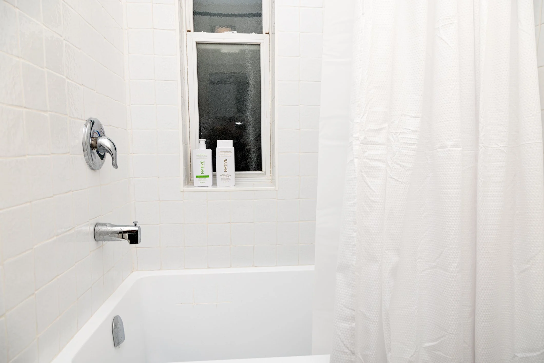 A bathroom shower area with white tiles, a small frosted window, two bottles of shampoo or body wash, a shower faucet, and a white shower curtain.