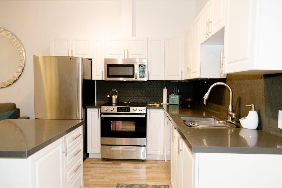Modern kitchen with white cabinets, stainless steel refrigerator, microwave, oven, and black backsplash. Countertop with sink, soap dispenser, and a few items like a wine box and paper towel roll.
