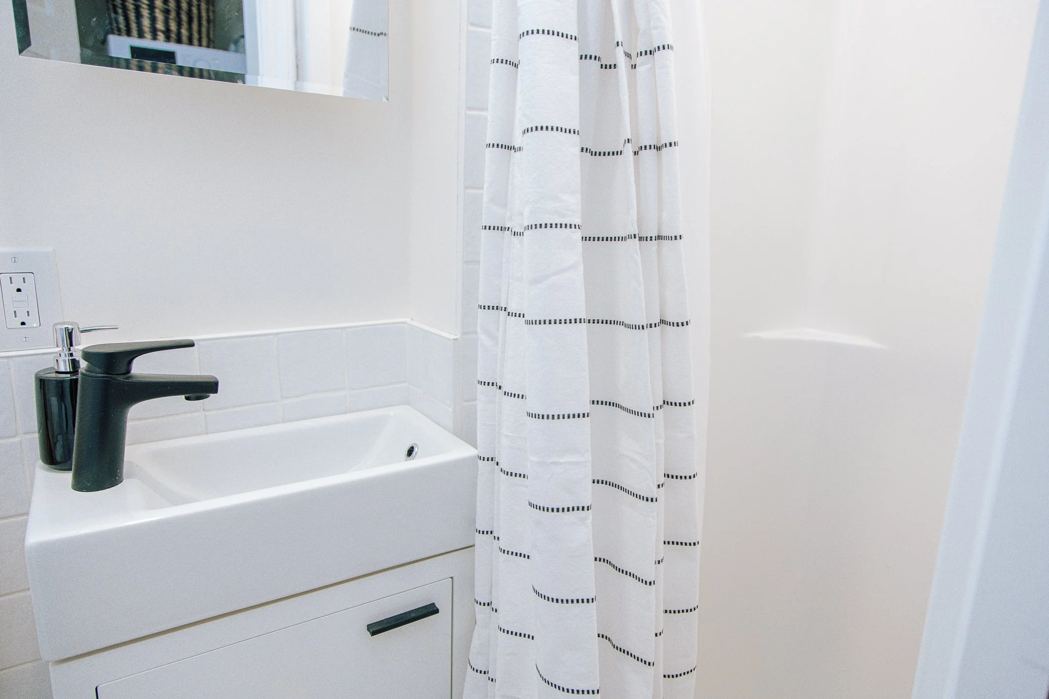 Bathroom with a white sink, black faucet, mirror, power outlet, and a white shower curtain with black dashed stripes.