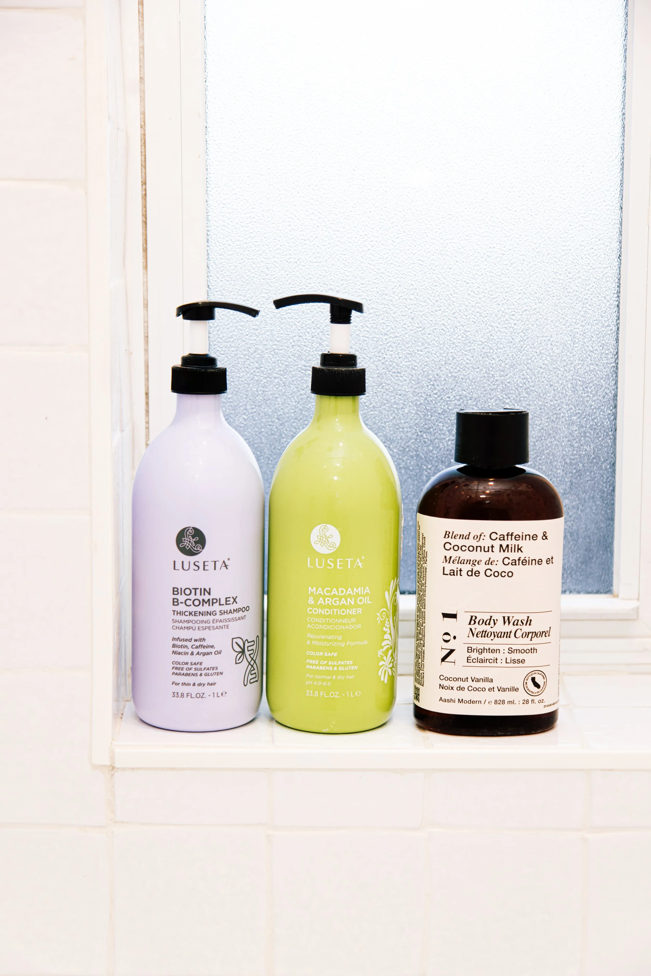 Three bottles of hair and body care products are displayed on a white shelf. The first bottle on the left is white with a pump labeled 'LUSETÀ Biotin B-Complex Shampoo.' The middle bottle is yellow with a pump labeled 'LUSETÀ Macadamia & Argan Oil Co