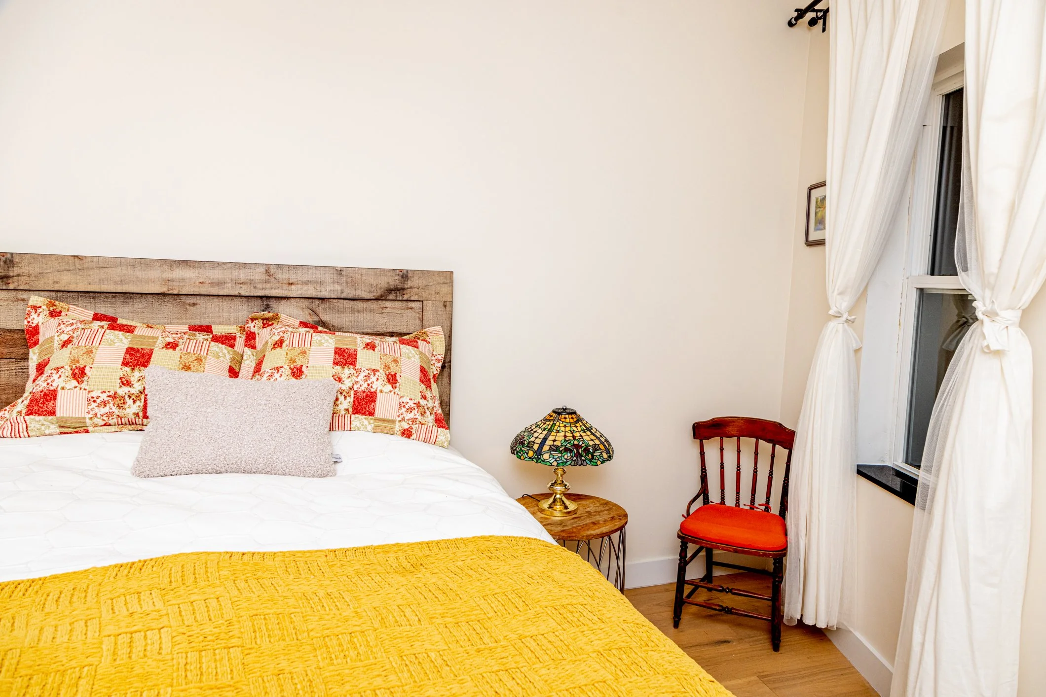 A bedroom corner with a bed, featuring a patchwork quilt and pillows, a Tiffany-style stained glass lamp on a wooden nightstand, a wooden chair with an orange cushion, and a window with white curtains.