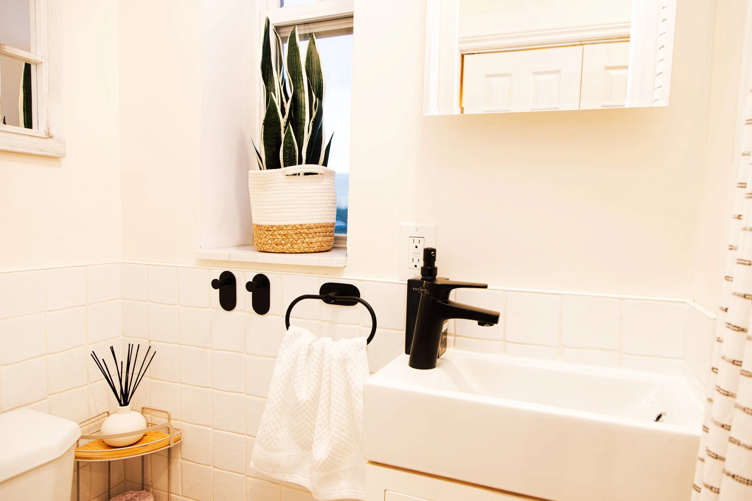 A bathroom with a white tile wall, a black faucet, a mirror, and a window with blinds. There is a white towel hanging on a black towel ring, and a small wooden side table with a decorative vase and reed diffuser. A plant in a woven basket is on the w