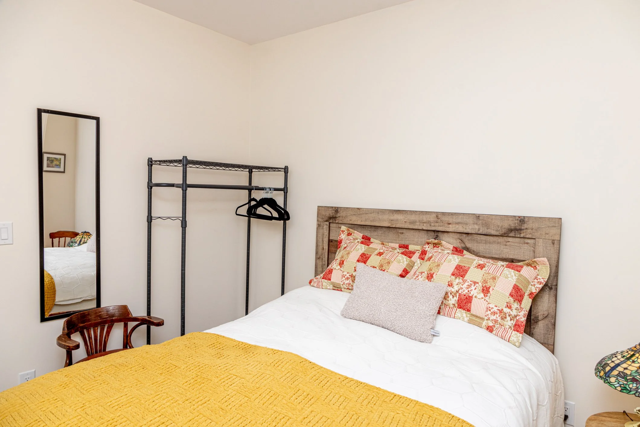 A bedroom with a bed that has a patchwork quilt, two pillows, and a rustic wooden headboard. There is a yellow bedspread in the foreground, a full-length mirror on the wall, and a black metal clothing rack with hangers. A wooden chair is partially vi