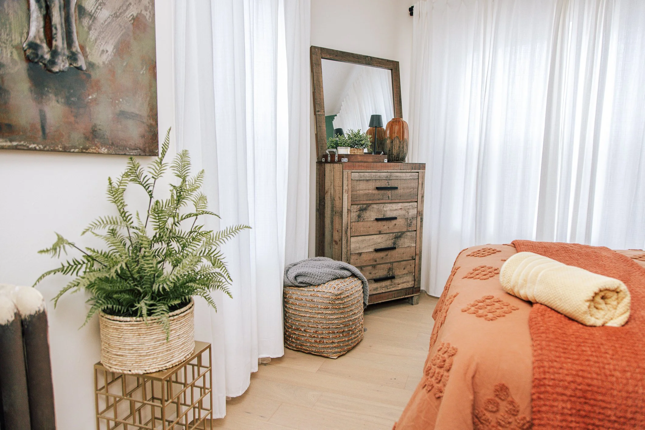 A cozy bedroom corner with a wooden dresser topped with vases, plants, and a lamp, contrasted with white curtains, a green potted fern on a small table, and an orange bedspread with rolled towels.