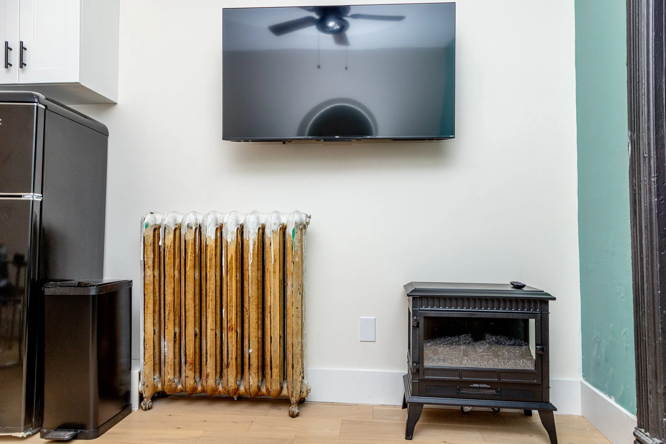 Interior of a room with a wall-mounted flat-screen TV, an old radiator with peeling paint, a black small table with a remote on top, a black cabinet, and part of a white cabinet visible on the left. The wall on the right has a teal accent. The floor 