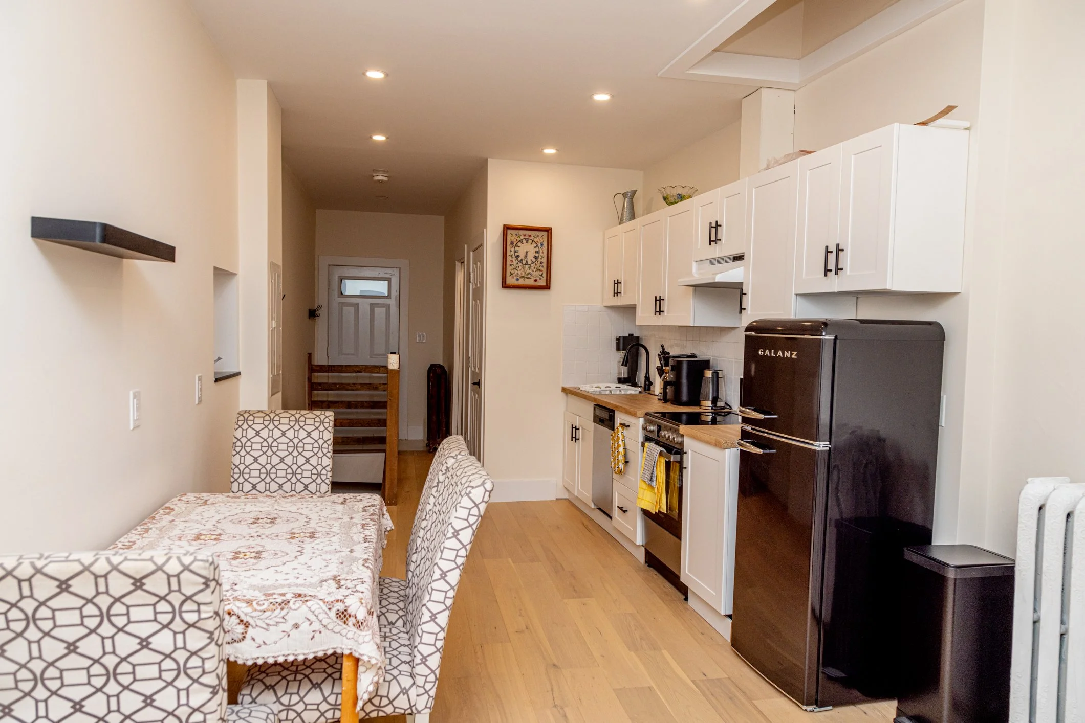 A kitchen and dining area with white cabinets, black appliances, a wooden countertop, a dining table with patterned chairs, and a hallway leading to a front door.