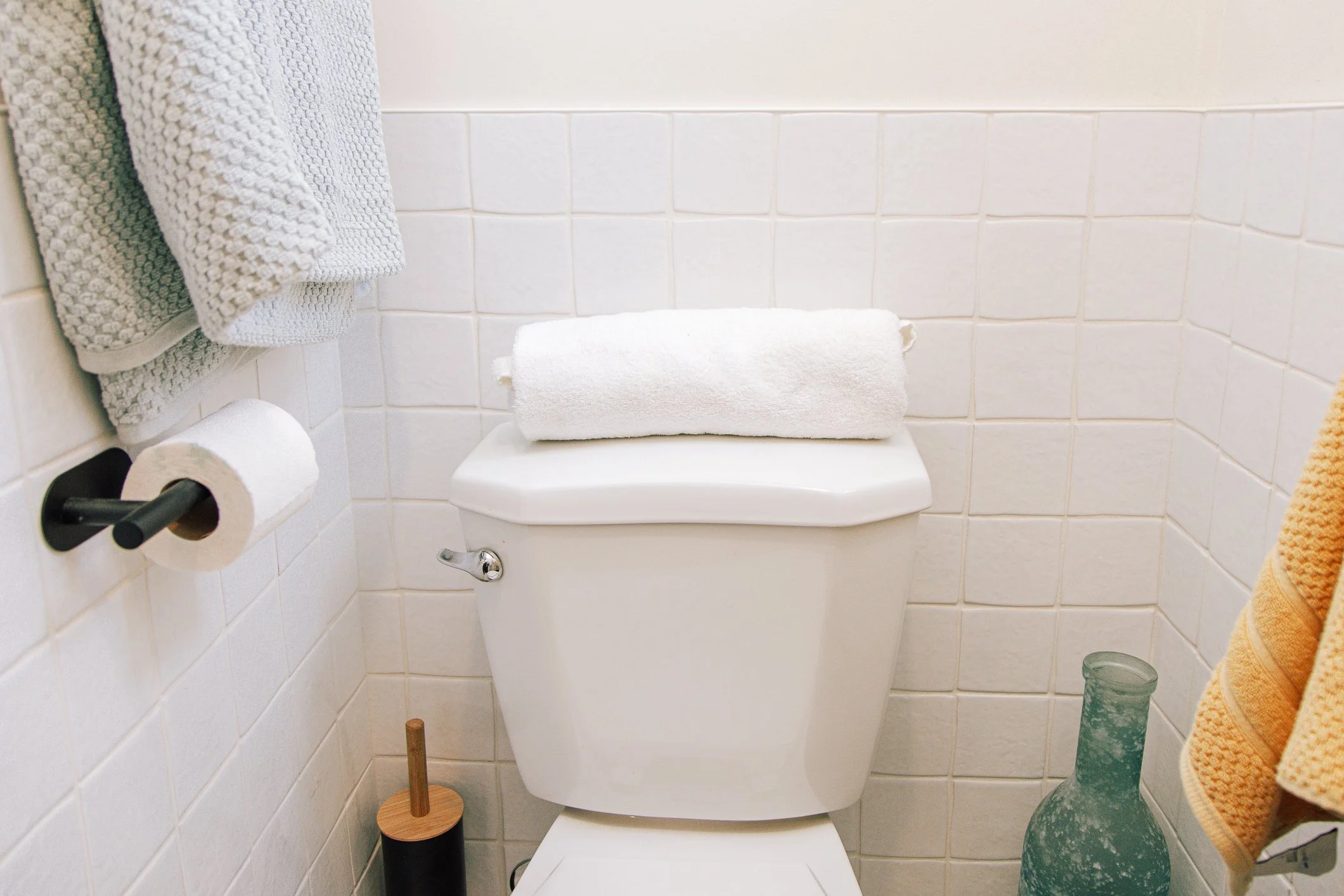 A white toilet with a rolled white towel on the lid, surrounded by beige and white wall tiles. There are two towels, one gray and one green, hanging on the wall, and a toilet paper roll on a black holder on the left side. A wooden toilet brush with a