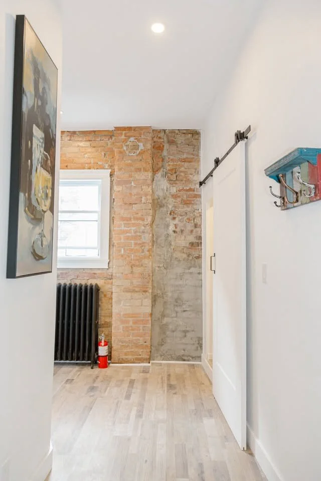 Interior of a room with a brick wall, a window, a black radiator, a fire extinguisher, a white sliding barn door with a black track, and a colorful wall hook.