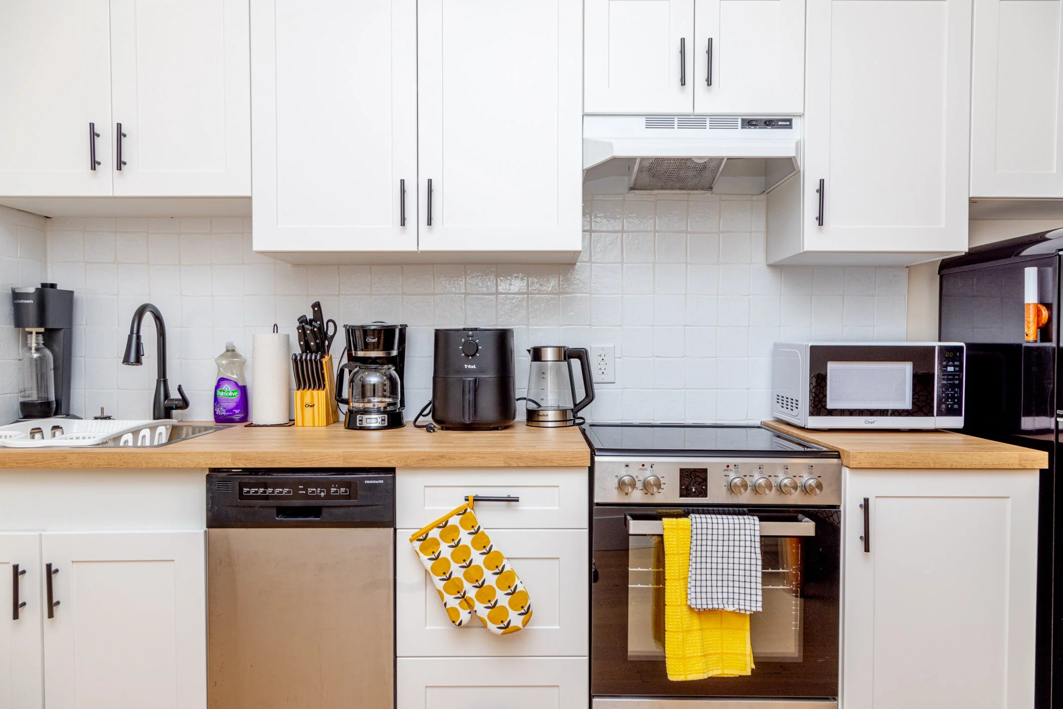 Kitchen with white cabinets and a wooden countertop, featuring various appliances such as a microwave, toaster, coffee maker, kettle, and microwave oven. There are dish towels hanging from cabinet handles and oven, and a spray bottle and knife block 