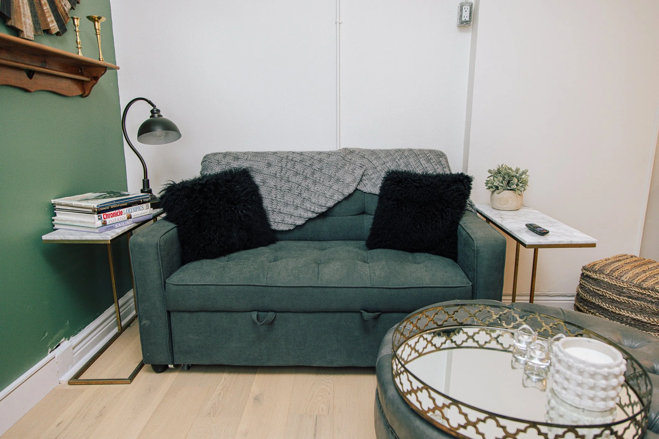 A cozy living room corner with a green sofa adorned with black furry pillows, gray knitted blanket, a side table with a stack of magazines, a black desk lamp, a potted plant, a remote, and a decorative round glass table with candles and ornaments. Th