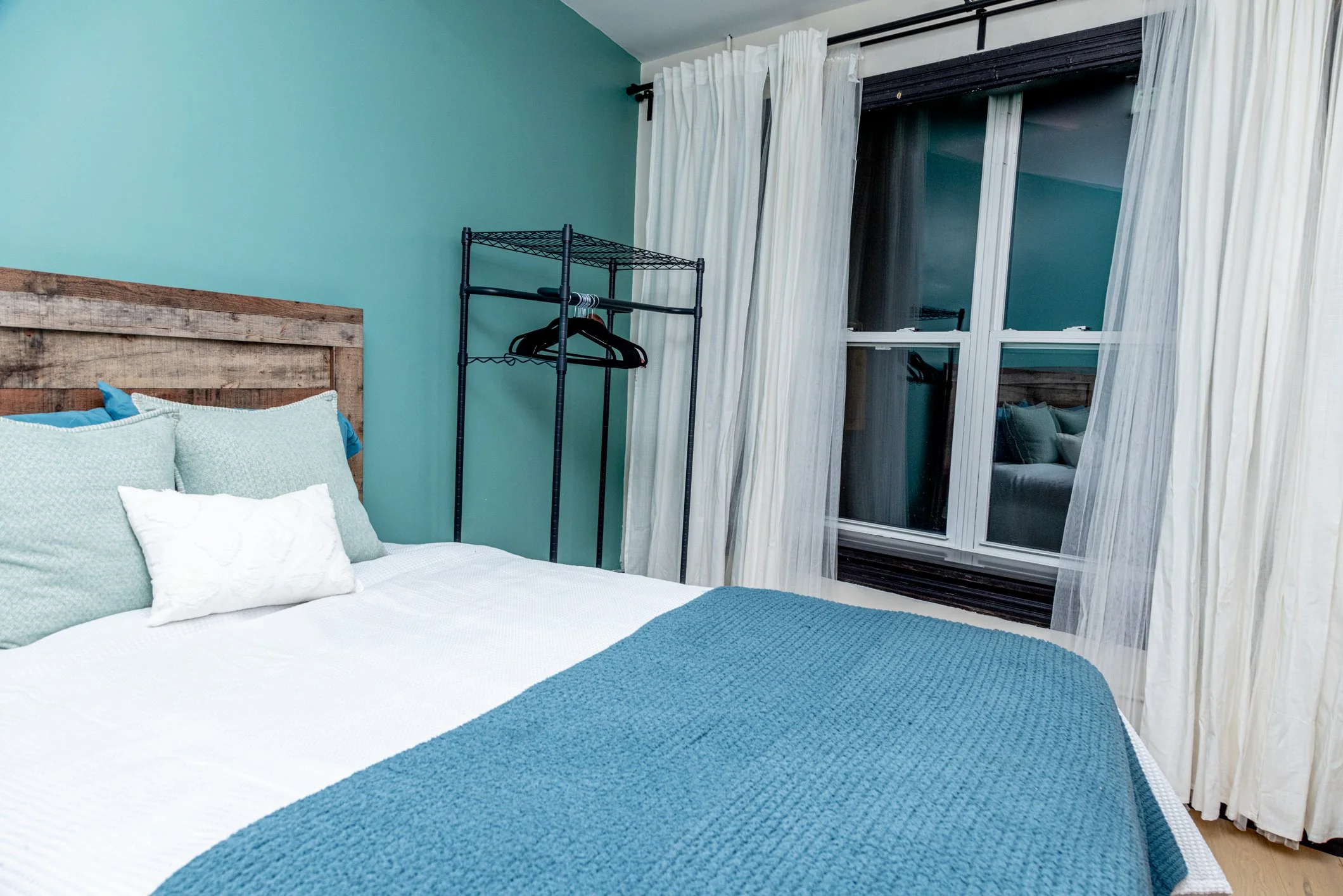 A cozy bedroom with a bed that has white and blue bedding and pillows. A black metal clothing rack with few hangers is positioned near a large window with white curtains. The walls are painted teal, and a reflection of the bed can be seen in the wind
