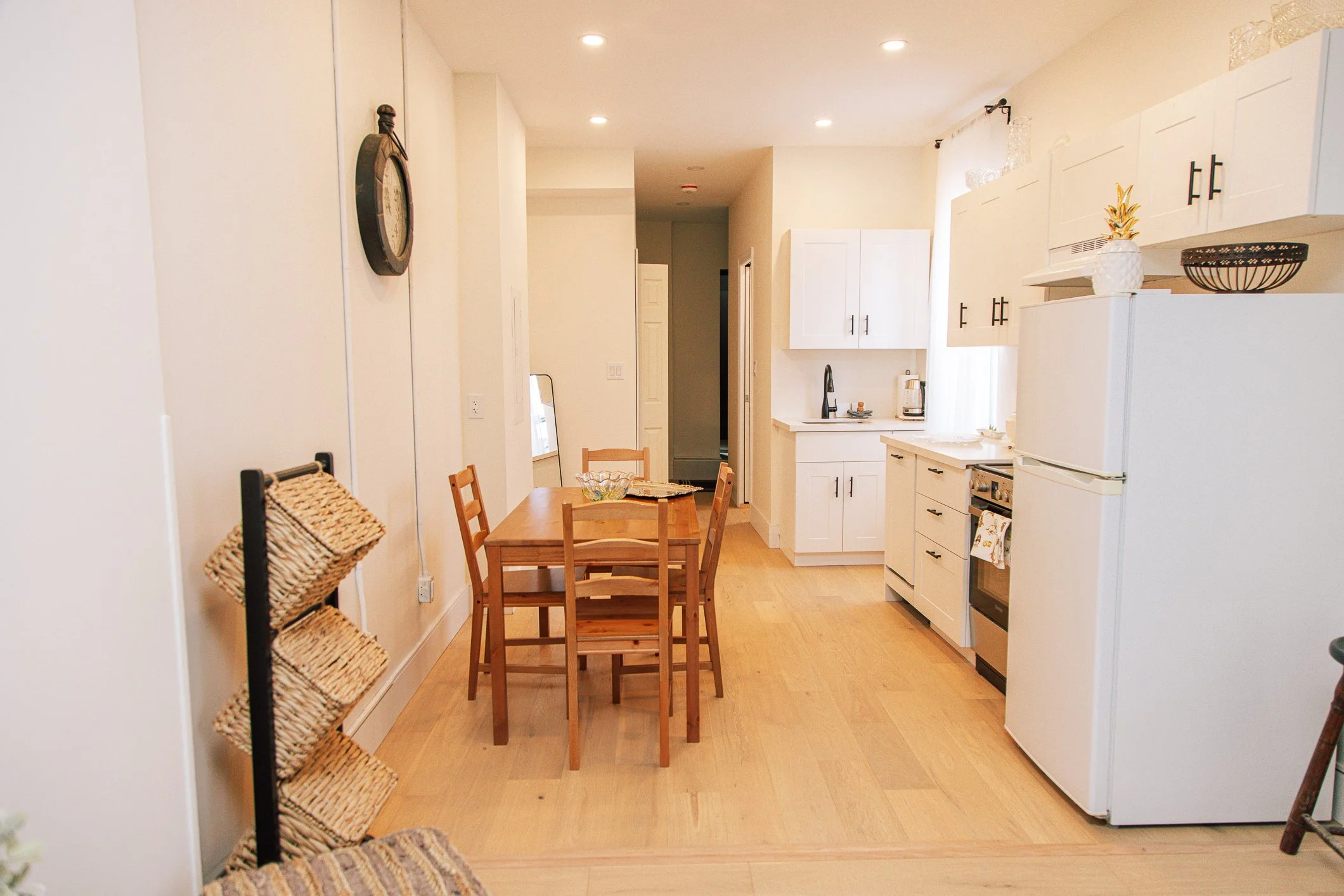 A cozy kitchen and dining area with white cabinets, a small wooden table with four chairs, a white refrigerator, and a black stove. Decor includes a pineapple-shaped container, a black bowl, and glass jars on top of the cabinets. Soft natural light f