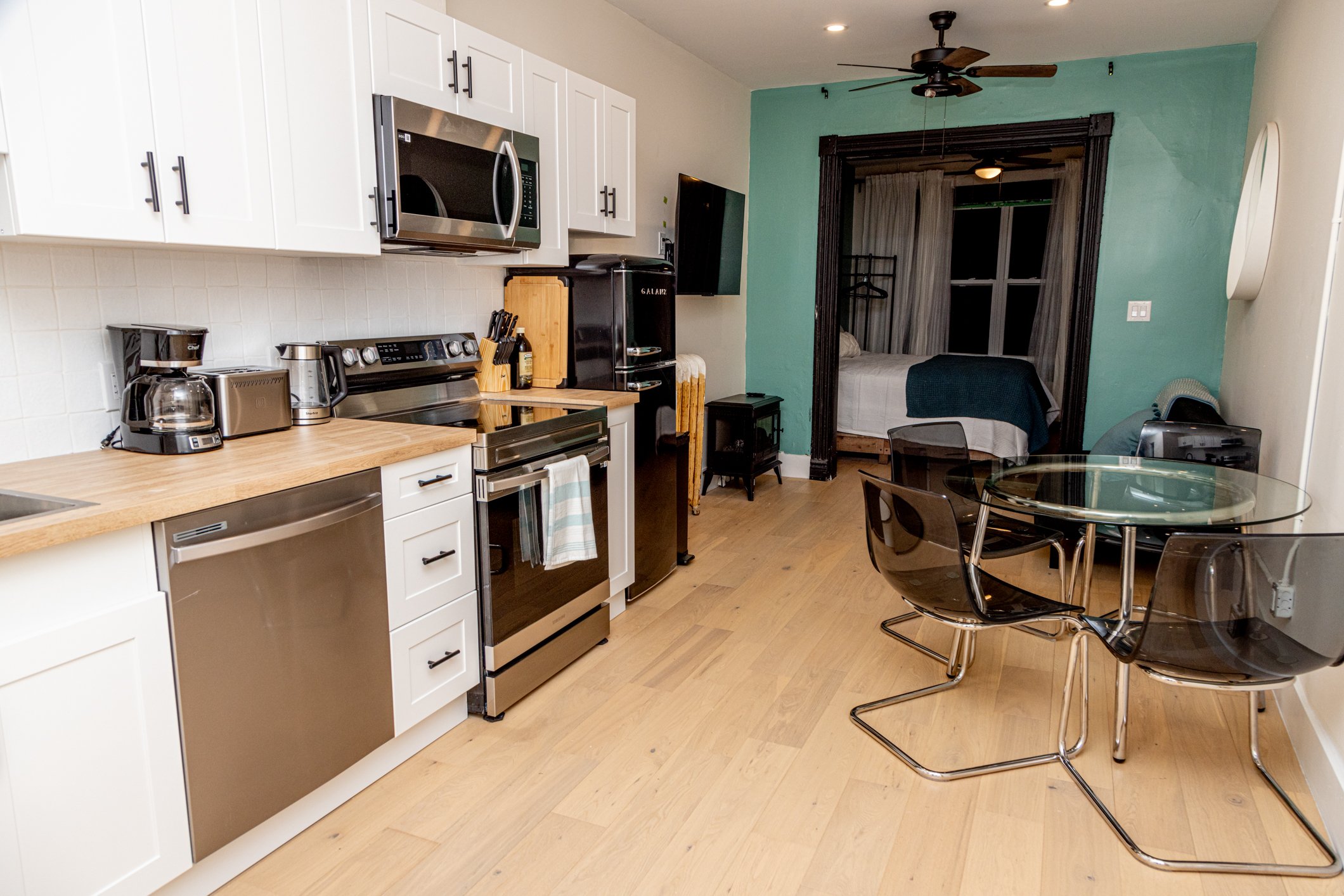 A kitchen with white cabinets, a stainless steel microwave, stove, and dishwasher, a coffee maker, toaster, and knife block on the wooden countertop. Adjacent is a dining area with a glass table and black chairs. In the background, a bedroom with a b
