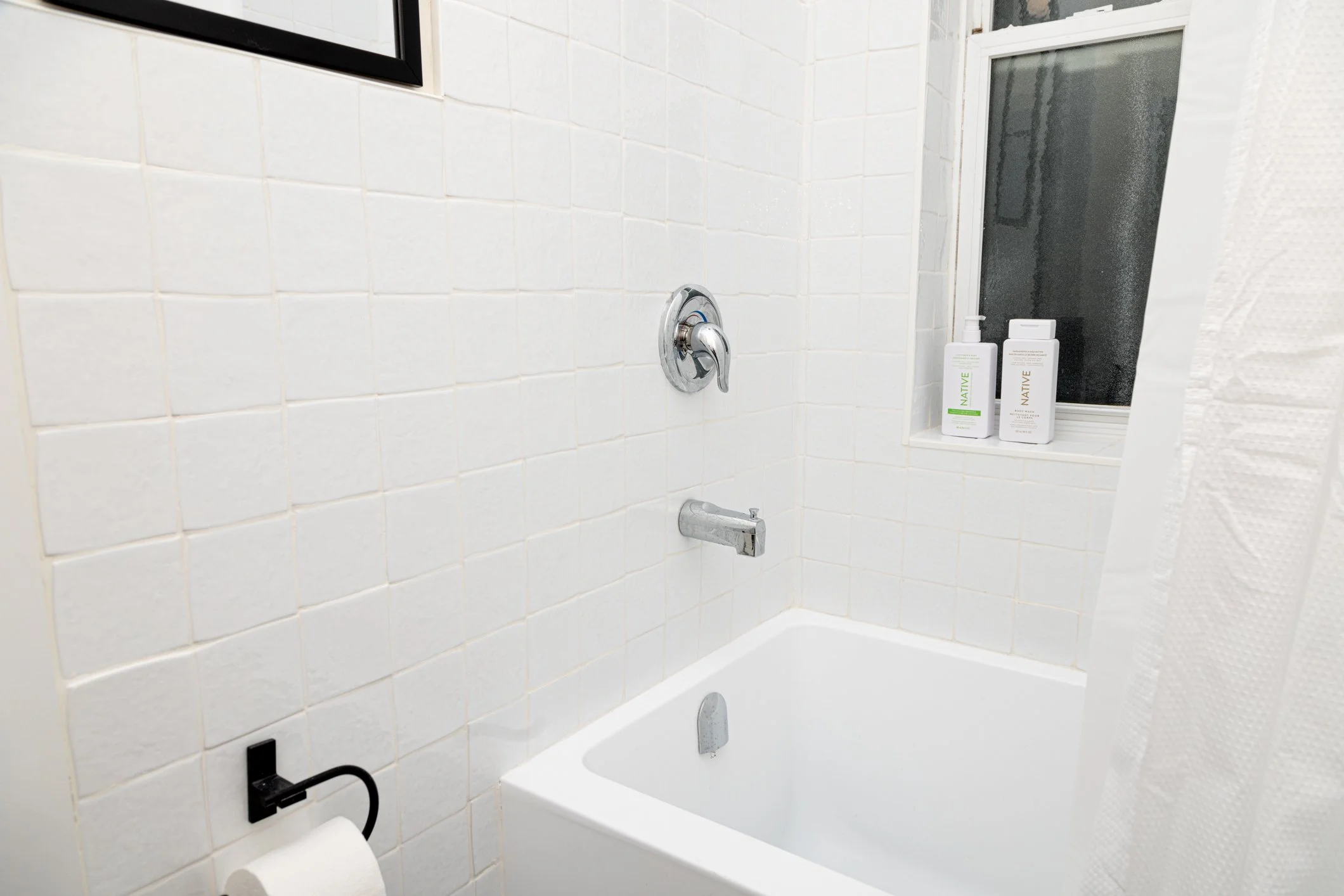 Bathroom with white tiled wall, a bathtub, a silver faucet, a black toilet paper holder, and two bottles of Native brand body wash on the window sill.