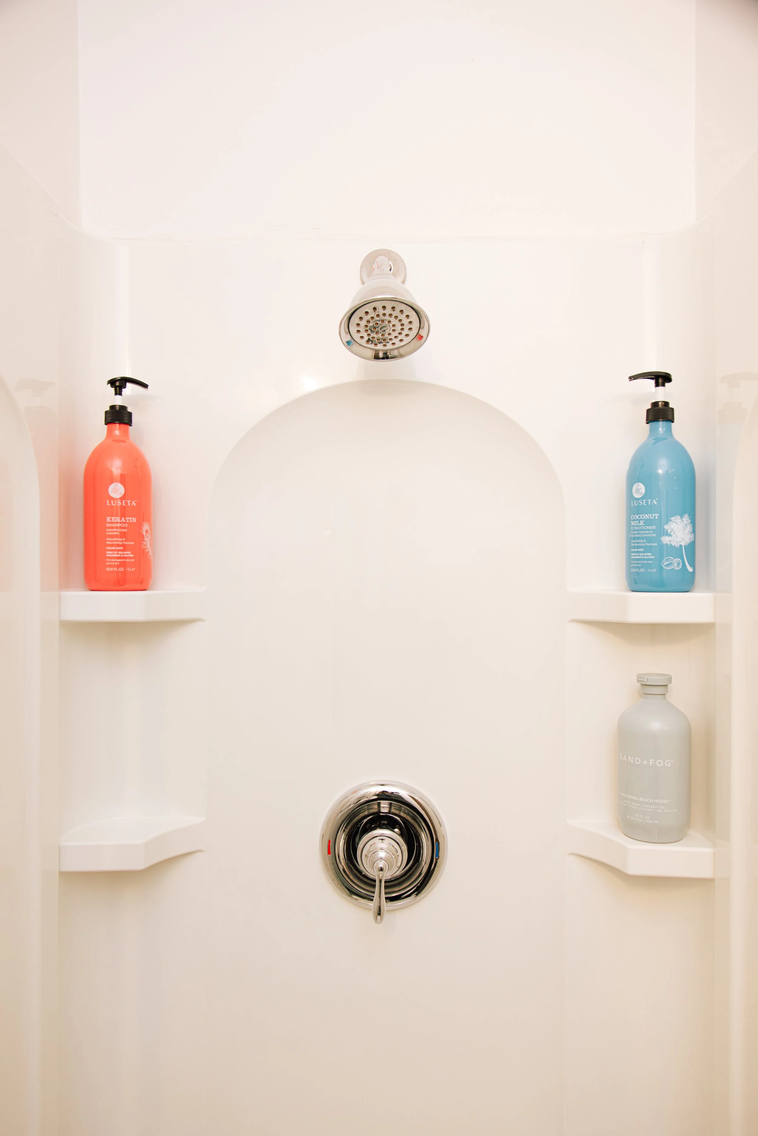 An overhead view of a white shower stall with a round showerhead, a single stream of water, and two built-in shelves holding bottles of shampoo, conditioner, and body wash.