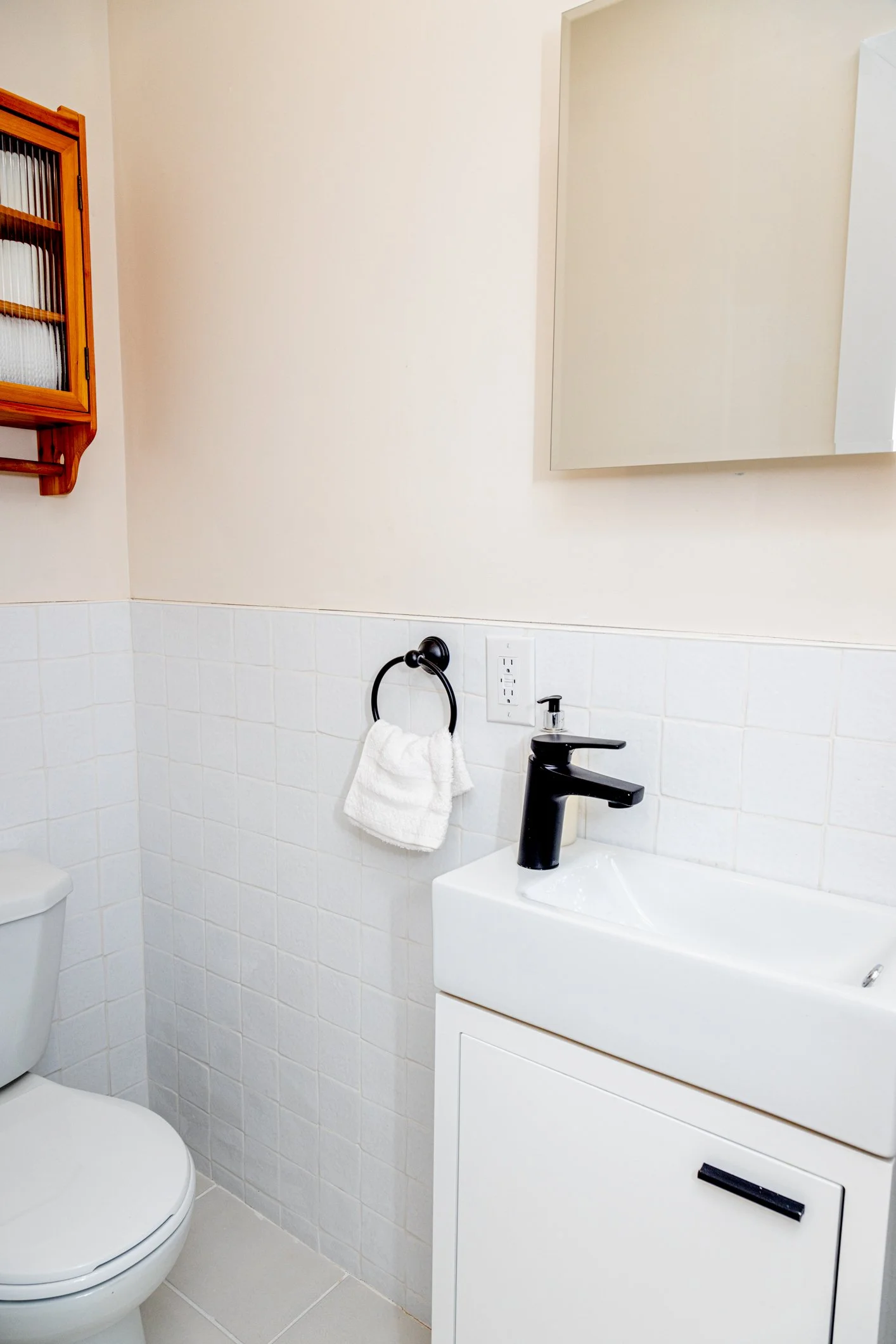 Bathroom with white tiled wall, black faucet, mirror, towel ring with a towel, and a wooden cabinet.