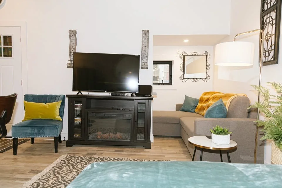 Living room with a flat-screen TV on a black cabinet, a blue velvet armchair with a yellow pillow, a beige sectional sofa with teal pillows and a yellow throw, a round side table with a plant, a tall white floor lamp, and decorative wall mirrors.