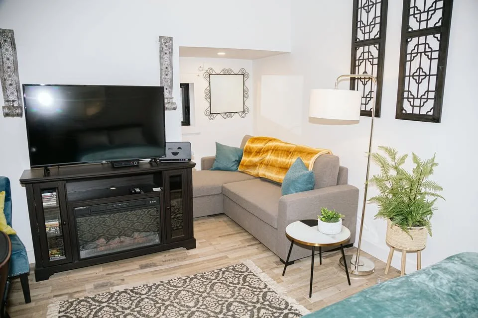 Living room with a large flat-screen TV on a black cabinet, a beige sofa with blue pillows and an orange throw blanket, a white round side table with a plant, a tall floor lamp, and decorative black window panels on the wall.