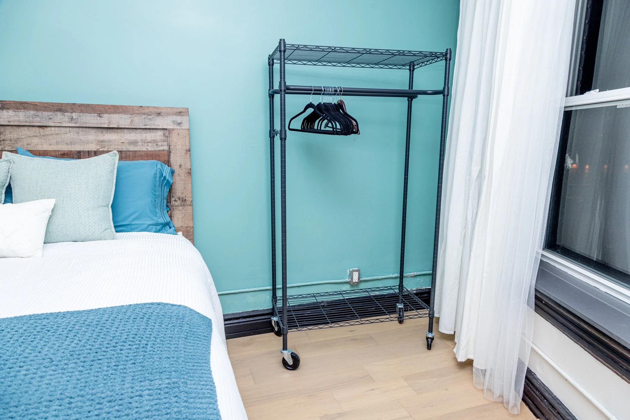 A black metal rolling clothing rack with a horizontal bar and a top shelf is positioned next to a bed with a rustic wooden headboard, blue and white bedding, and pillows in a bedroom with light green walls, white curtains, and a large window.