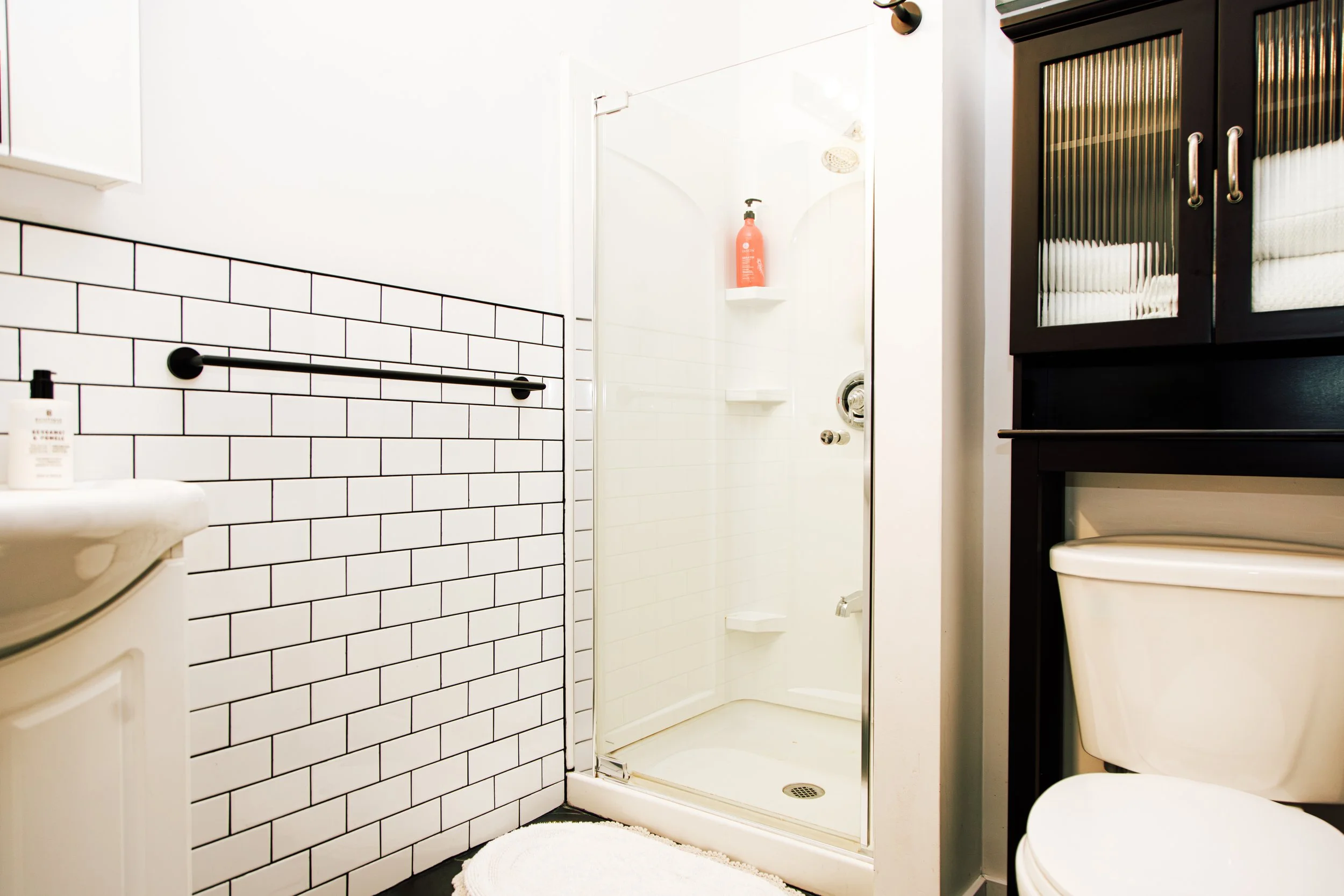 A modern bathroom with a white subway tile wall, a white sink, a black towel bar, a shower with glass door, a dark wooden cabinet, a white toilet, and a white round rug.