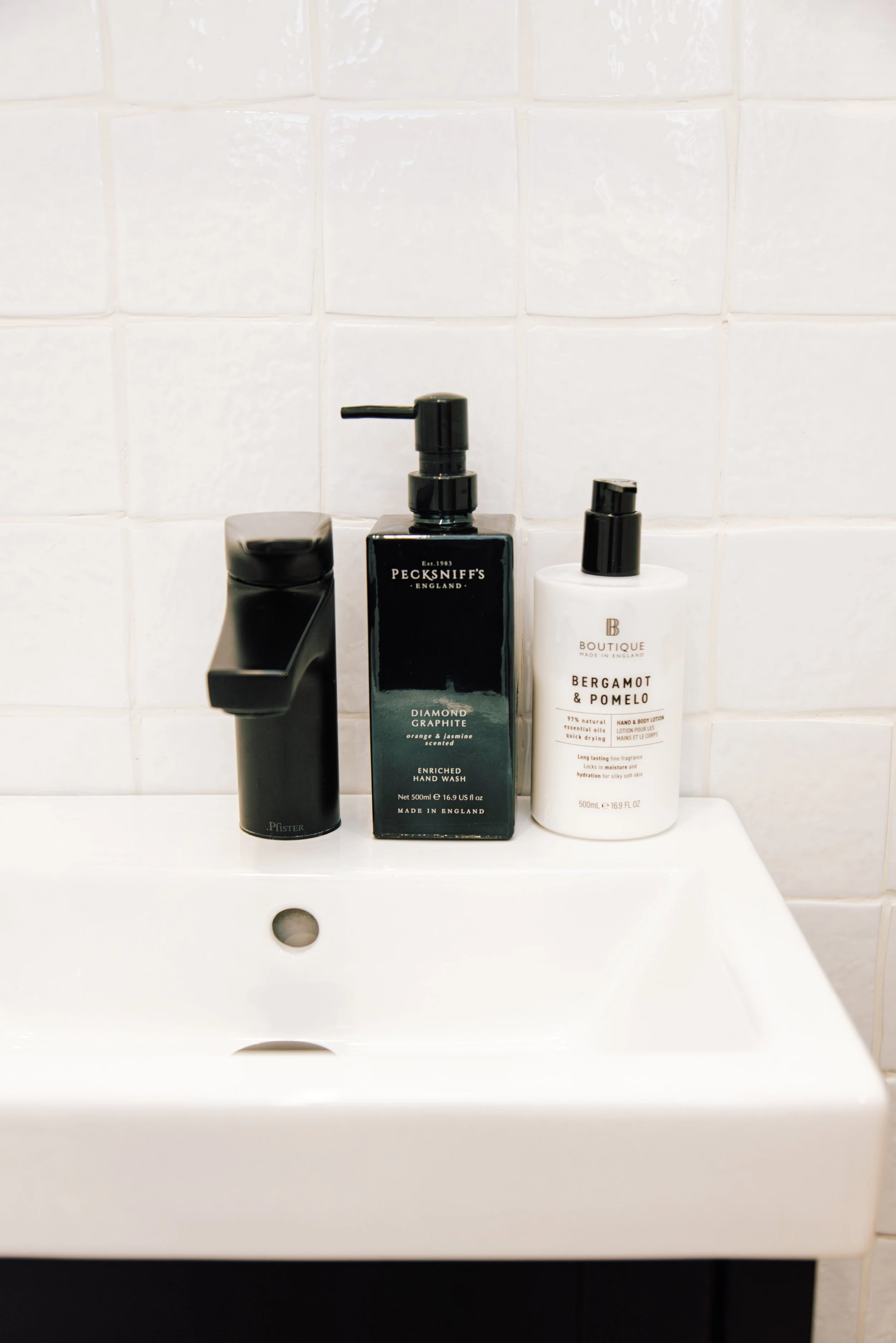 A white bathroom sink with a black faucet, with three bottles of personal care products on the sink. The bottles include a black pump dispenser labeled "PECKSNIFF'S," a white bottle labeled "BERGAMOT & POMELO," and a small black and white container. 