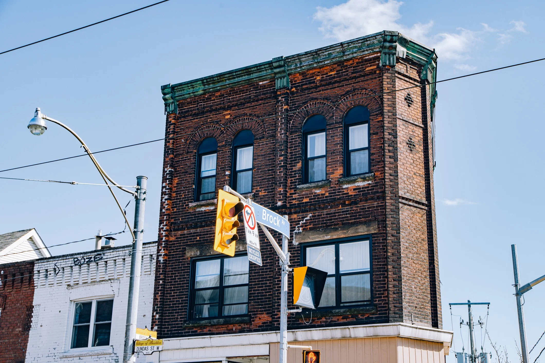 An old brick building with four tall windows on the top floor, some with white curtains. The corner of the building is visible. There are electrical wires crossing in front of the building, and a street sign indicating Brock Avenue and Dundas Street West. A traffic light and street lamps are also visible against a clear blue sky.