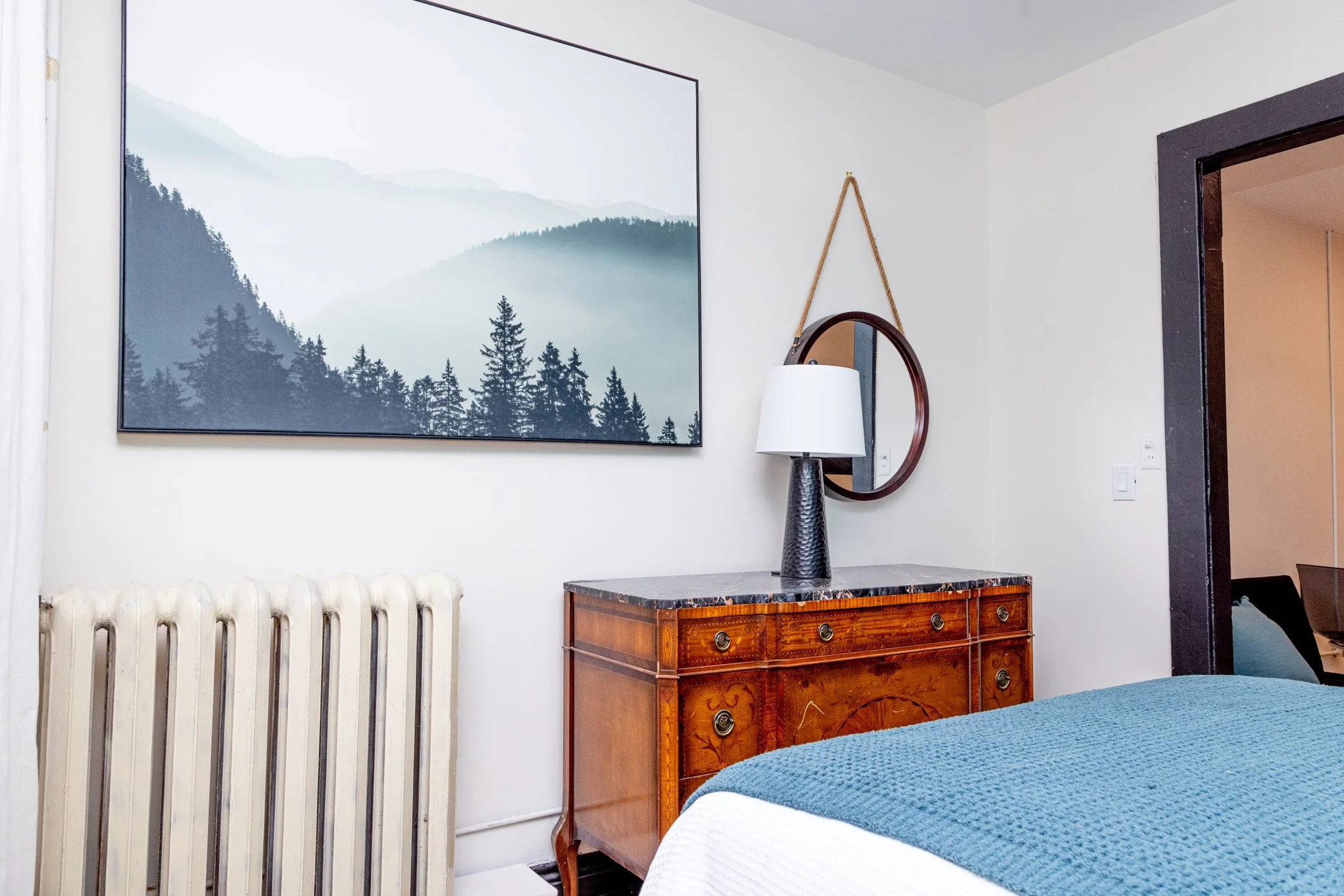 A bedroom corner with a white radiator beneath a mountain landscape painting. There's a wooden dresser with a tall black textured vase, a white lampshade, and a hanging mirror. Part of a bed with a blue textured blanket is visible in the foreground.